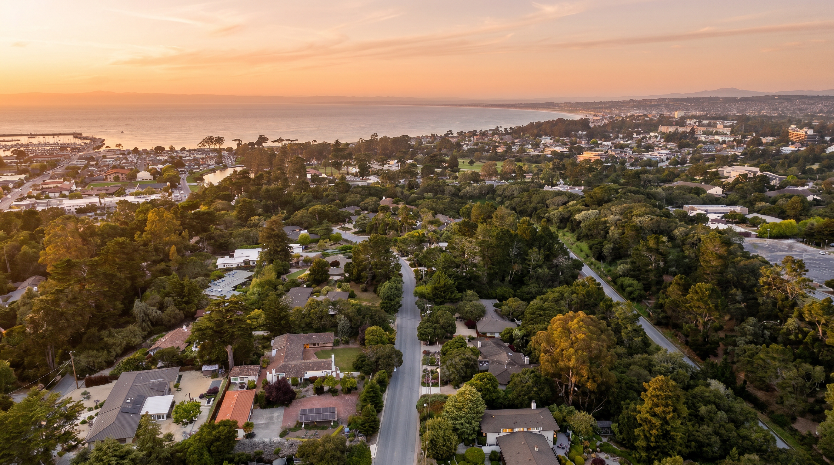 This high-angle aerial perspective captures a coastal residential neighborhood nestled among dense, mature trees. The scene features a winding road leading toward a scenic bay, with a clear blue sky and distant mountains providing a picturesque backdrop. The composition highlights the lush, green landscape and the proximity of the homes to the ocean, emphasizing a tranquil and desirable coastal lifestyle.