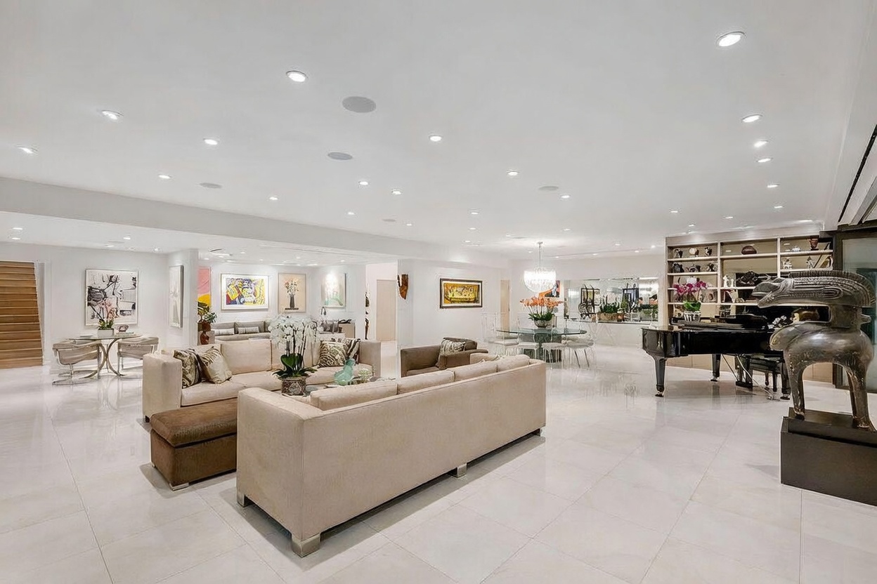 This expansive, open-concept living area features polished light-colored flooring and a neutral color palette, creating a bright and airy atmosphere. The space is furnished with multiple plush sofas, a grand piano, and a dining area, all illuminated by numerous recessed ceiling lights and a central chandelier. A large, abstract sculpture stands in the foreground, adding a sophisticated artistic touch to this modern, high-end interior.