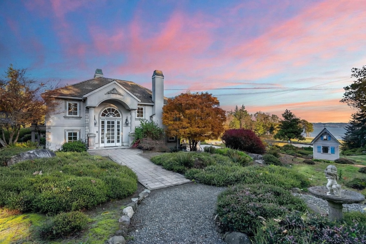 This elegant two-story residence features a classic architectural style with a prominent arched entryway and a stone-paved walkway leading to the front door. The home is set against a vibrant sunset sky, with a lush, landscaped front yard, a small decorative playhouse, and a stone birdbath in the foreground. The property offers a picturesque view of the water in the distance, creating a serene and inviting atmosphere.