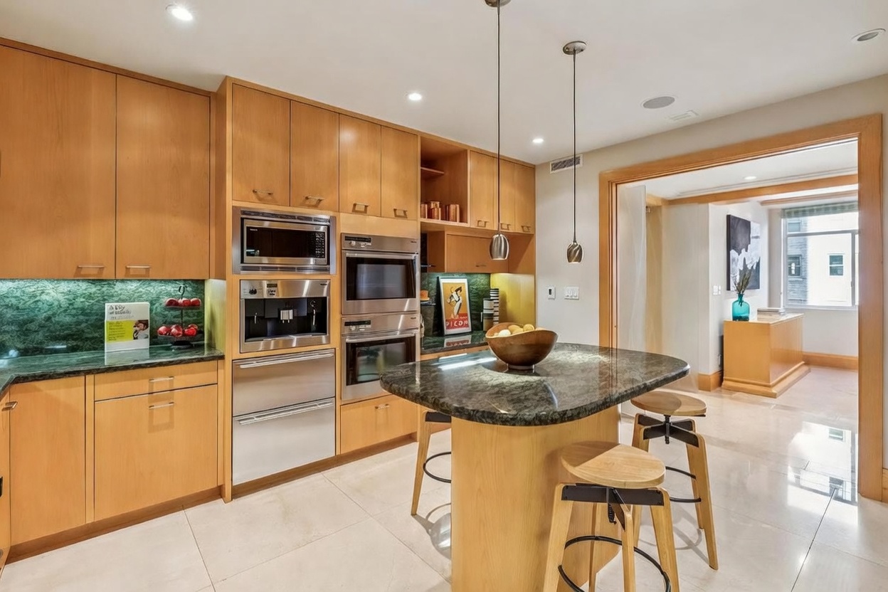 This modern kitchen features light wood cabinetry, integrated stainless steel appliances, and a central island topped with dark stone. The space is illuminated by recessed lighting and two pendant lights hanging over the island, which is accompanied by two wooden bar stools. A doorway leads into an adjacent room, creating an open and connected floor plan with a bright, neutral aesthetic.