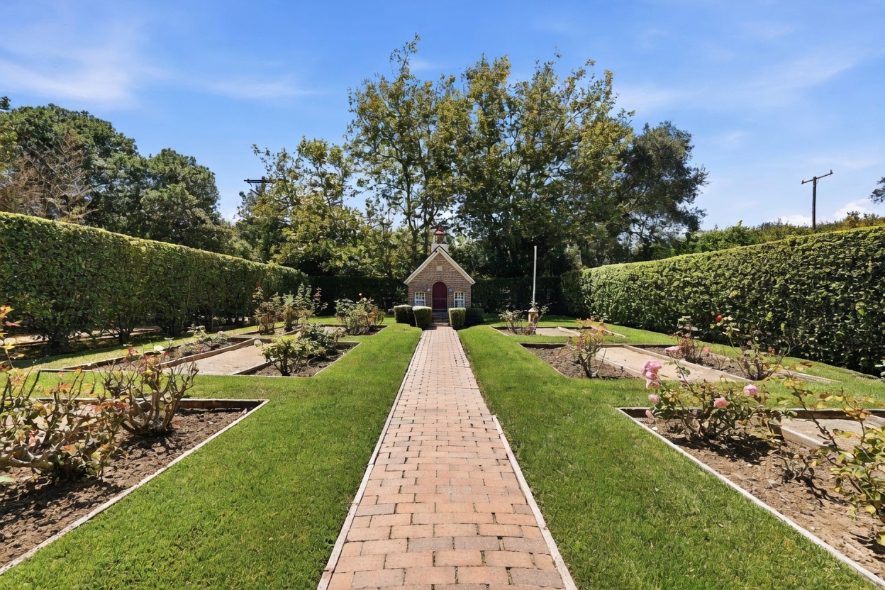This formal garden features a symmetrical layout with a central brick pathway leading toward a charming, small brick structure with a red door. The garden is framed by tall, manicured hedges and includes several planting beds filled with rose bushes and other greenery. The scene is captured from a ground-level perspective, emphasizing the orderly and serene atmosphere of the outdoor space.