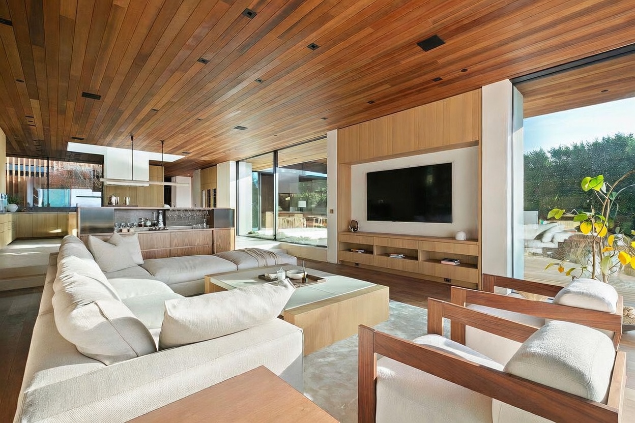 This sophisticated, open-concept living room features a warm, natural aesthetic with extensive wood-paneled ceilings and floor-to-ceiling glass walls that seamlessly blend indoor and outdoor living. The space is anchored by a plush, light-toned sectional sofa, a minimalist wooden coffee table, and a custom built-in media console surrounding a large flat-screen television. The perspective captures the airy, expansive layout, highlighting the high-end finishes and the tranquil view of the lush greenery outside.