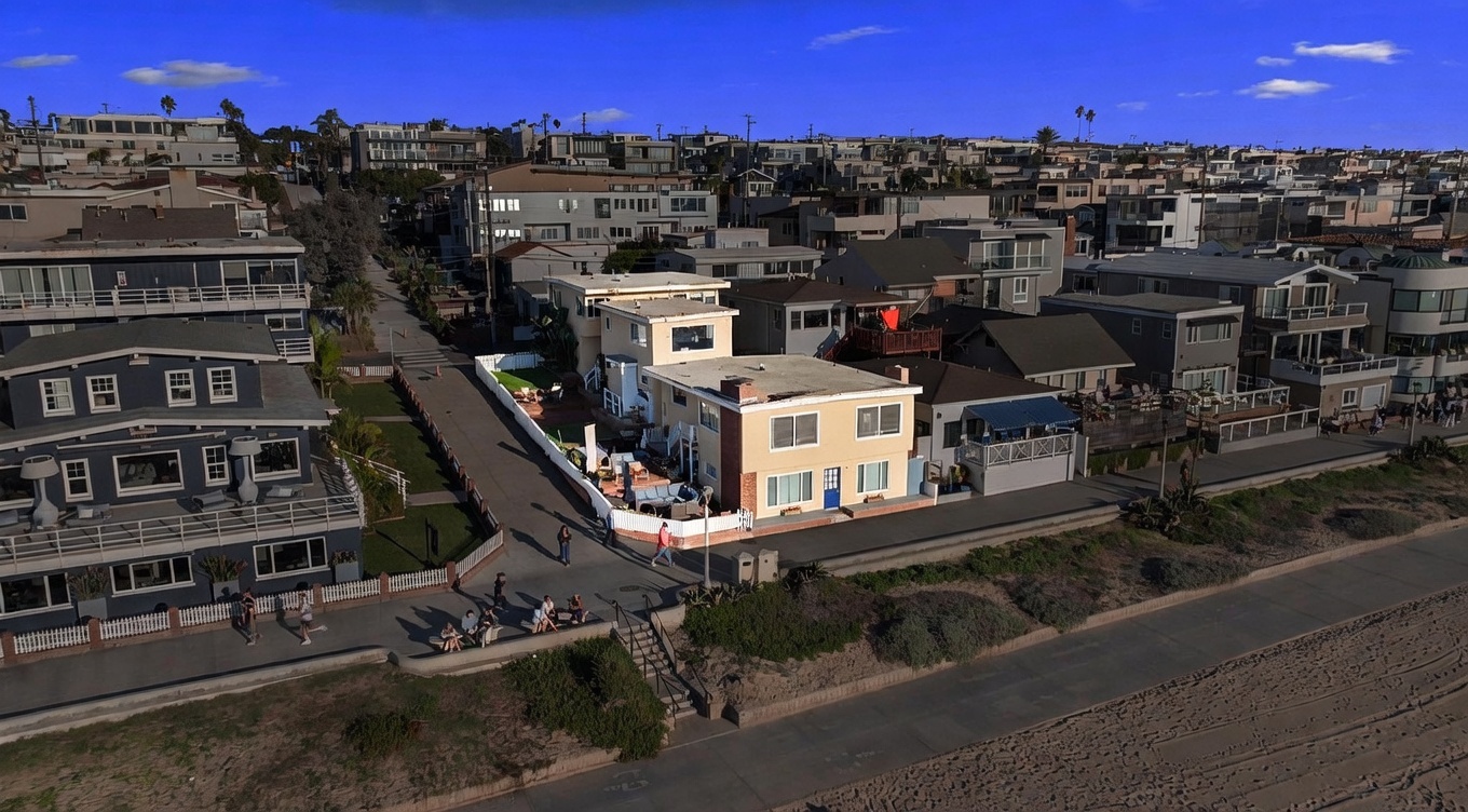 This high-angle aerial shot captures a coastal property situated directly adjacent to a sandy beach and a paved boardwalk. The primary residence is a light-colored, multi-story building with a flat roof, featuring a private fenced-in patio area with outdoor seating. The surrounding neighborhood consists of a dense collection of residential homes, showcasing a prime beachside location with clear blue skies above.