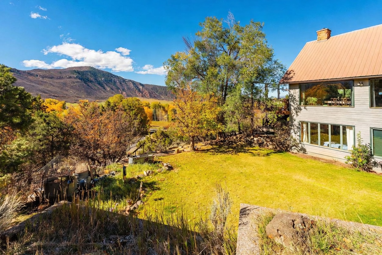 This exterior view captures the rear of a multi-story home with a metal roof, set against a backdrop of rolling hills and a vibrant autumn landscape. A spacious lawn leads toward a wooden deck attached to the house, which features large windows reflecting the surrounding trees and sky. The scene is framed by mature trees and a rustic stone foreground, emphasizing the property's peaceful, rural setting.