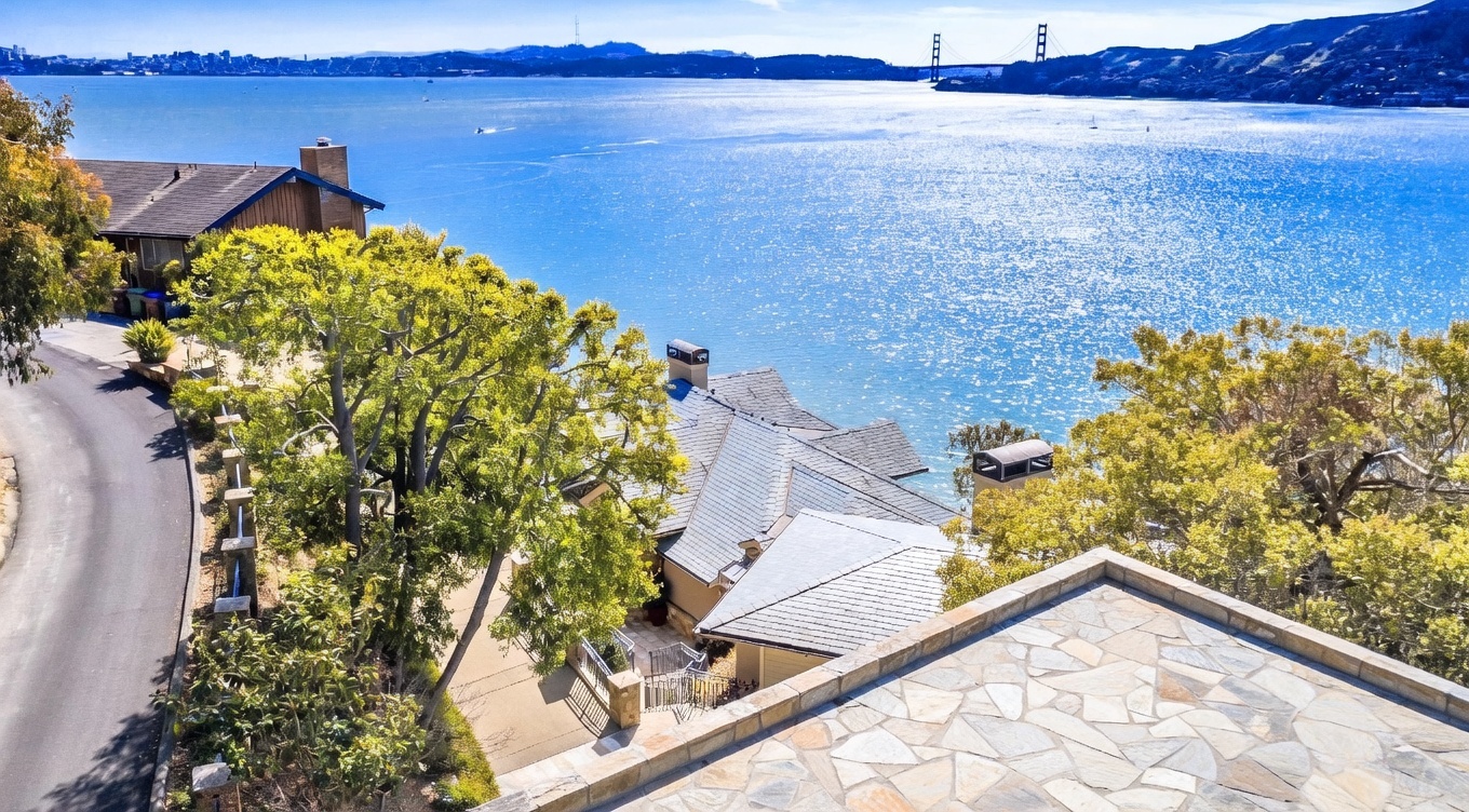 This high-angle aerial view captures a waterfront property nestled along a winding coastal road, showcasing its unique position overlooking a vast, sparkling bay. The home features a multi-gabled roofline with stone-clad chimneys and a paved driveway leading to a garage, all surrounded by mature trees. In the distance, the iconic Golden Gate Bridge is visible against the horizon, emphasizing the home's prime location and scenic surroundings.