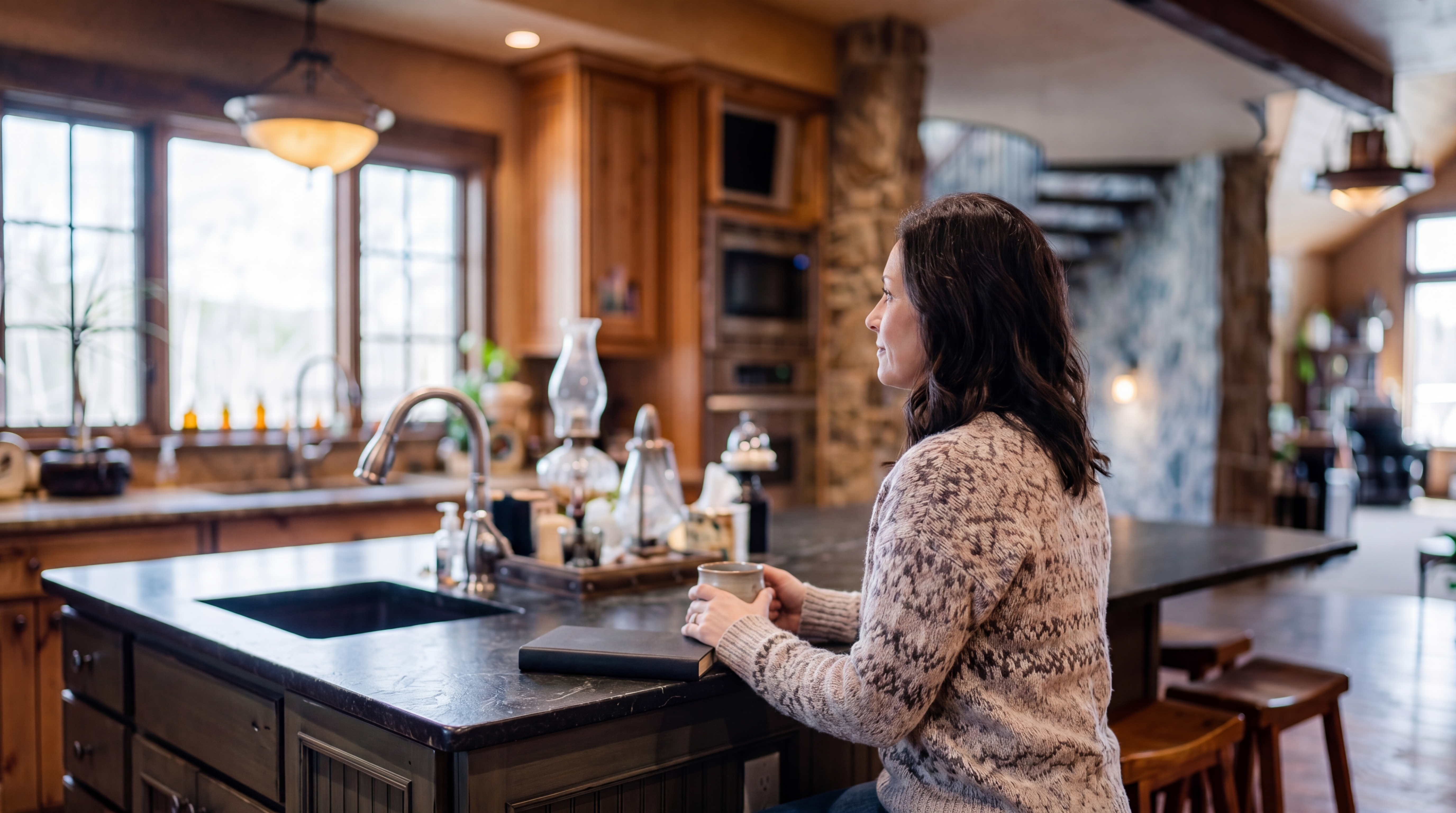 This rustic-style kitchen features warm wood cabinetry, a large central island with a dark stone countertop, and three wooden bar stools. The space is defined by exposed wooden ceiling beams and rich wood flooring, with a stone-clad wall leading toward a staircase in the background. The perspective captures the open-concept layout, highlighting the blend of natural textures and functional design elements.