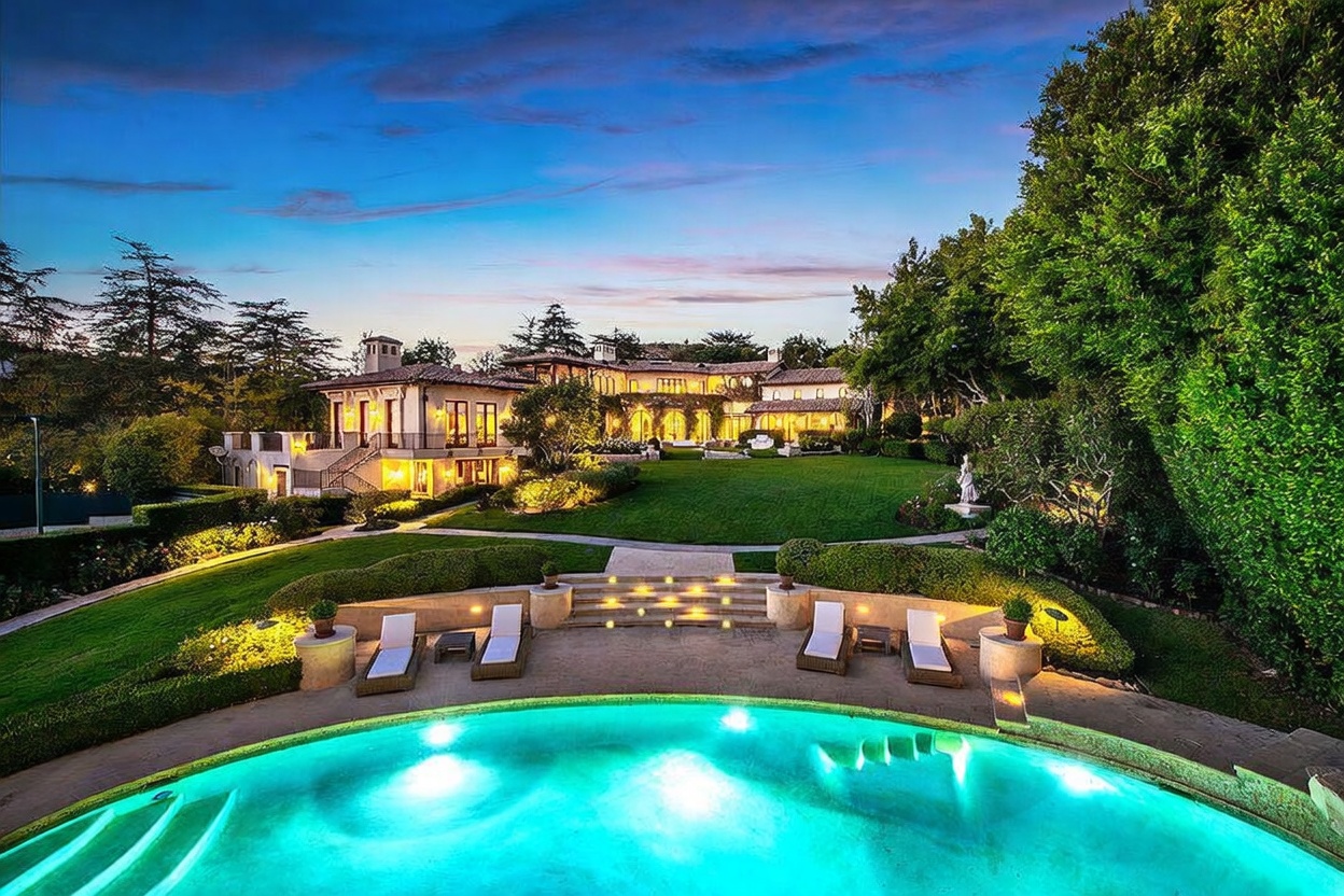 This expansive estate features a grand, multi-level backyard highlighted by a large, illuminated swimming pool in the foreground. Beyond the pool, a manicured lawn leads up to a sprawling, Mediterranean-style residence with warm exterior lighting and multiple balconies. The scene is captured at dusk, creating a serene and luxurious atmosphere that emphasizes the property's impressive scale and outdoor living potential.