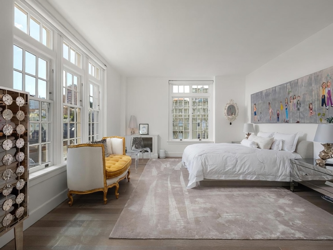 This bright and airy primary bedroom features a neutral color palette, light wood flooring, and a large area rug that anchors the space. A plush, gold-accented settee sits near a wall of tall windows, while a large, contemporary art piece hangs above the bed. The room is furnished with mirrored nightstands and lamps, creating a sophisticated and serene atmosphere.