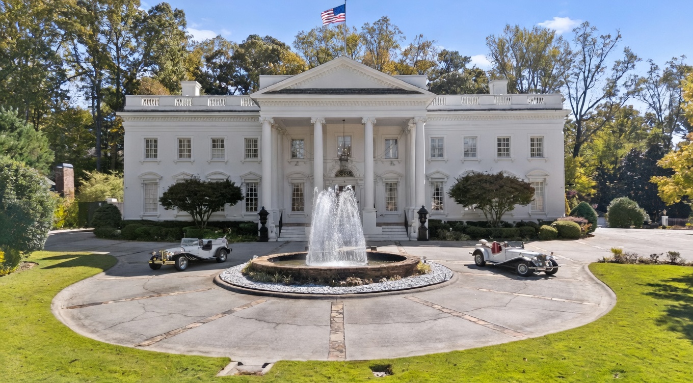 This grand, neoclassical-style estate features a symmetrical white facade with a prominent central portico supported by tall columns. A circular driveway surrounds a tiered stone fountain, flanked by two vintage-style convertible cars, creating a stately and formal arrival experience. The property is set against a backdrop of mature trees, emphasizing its expansive and private grounds.