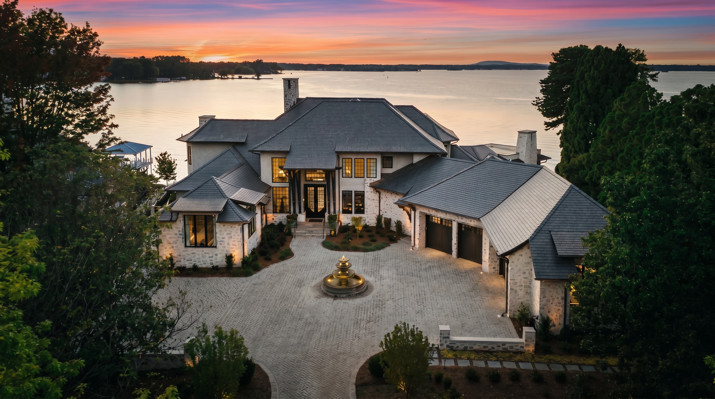 This stunning aerial perspective captures a luxurious waterfront estate at sunset, showcasing its expansive stone-clad facade and intricate slate-tiled roofline. A grand, circular brick-paved driveway leads to the main entrance, centered around an elegant tiered fountain. The property is nestled against a serene lake, with lush surrounding trees and a private dock visible, creating an atmosphere of sophisticated tranquility.