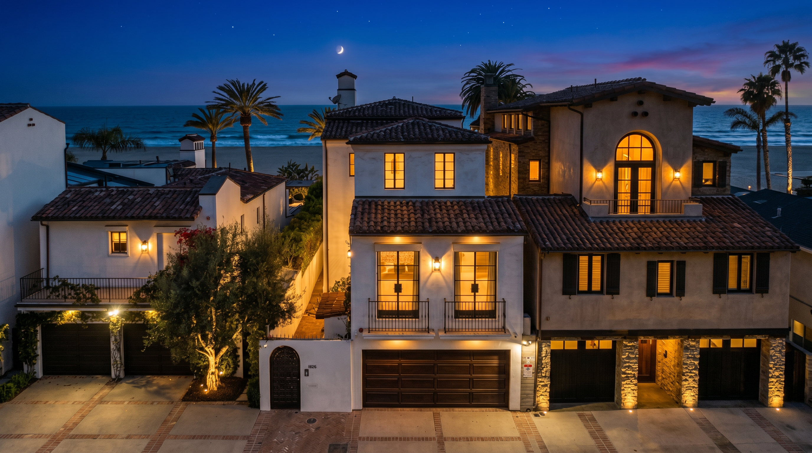 This striking aerial view captures a sophisticated multi-story beachfront estate at twilight, characterized by its classic Mediterranean architectural style. The property features rich brown tiled roofs, warm interior lighting glowing through numerous windows, and well-integrated multi-car garage spaces. Situated directly against the oceanfront, the home offers a seamless blend of luxurious living and coastal serenity under a crescent moon sky.