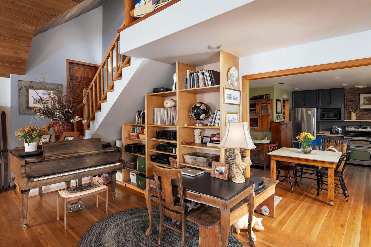 This open-concept living area features a warm, rustic aesthetic with light wood flooring and a prominent wooden staircase. A grand piano sits to the left, while a central shelving unit serves as a room divider, separating the living space from a dining area and kitchen visible in the background. The space is filled with personal touches, including a desk, a globe, and various decorative items, creating a cozy and lived-in atmosphere.