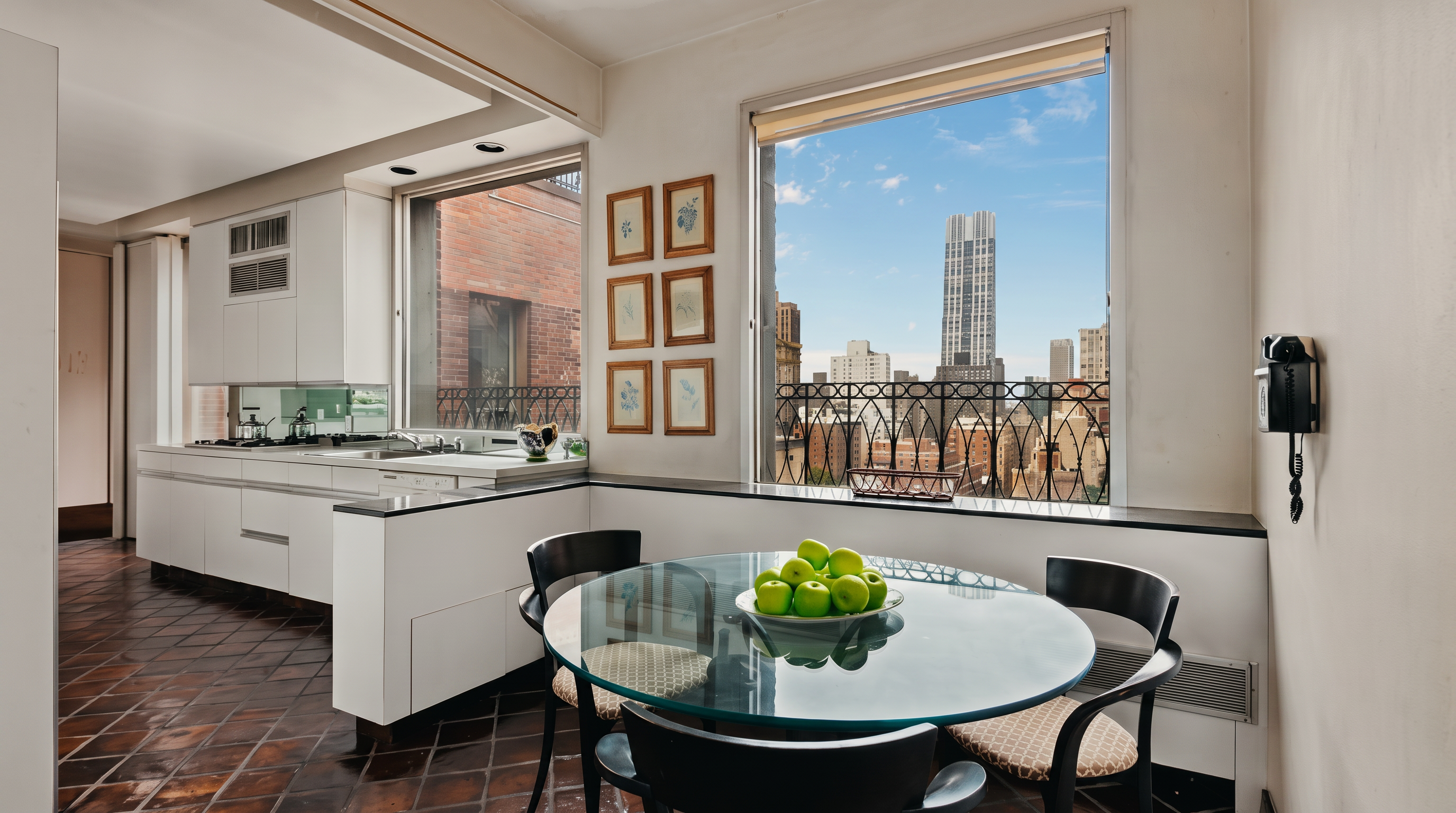 This bright dining area features a glass-topped circular table surrounded by four dark-framed chairs, positioned directly in front of a large window with an urban skyline view. The space is adjacent to a clean, white-cabinet kitchen with dark patterned tile flooring that extends throughout the room. A set of six framed artworks hangs on the wall, and a wall-mounted telephone adds a vintage touch to the corner of the space.