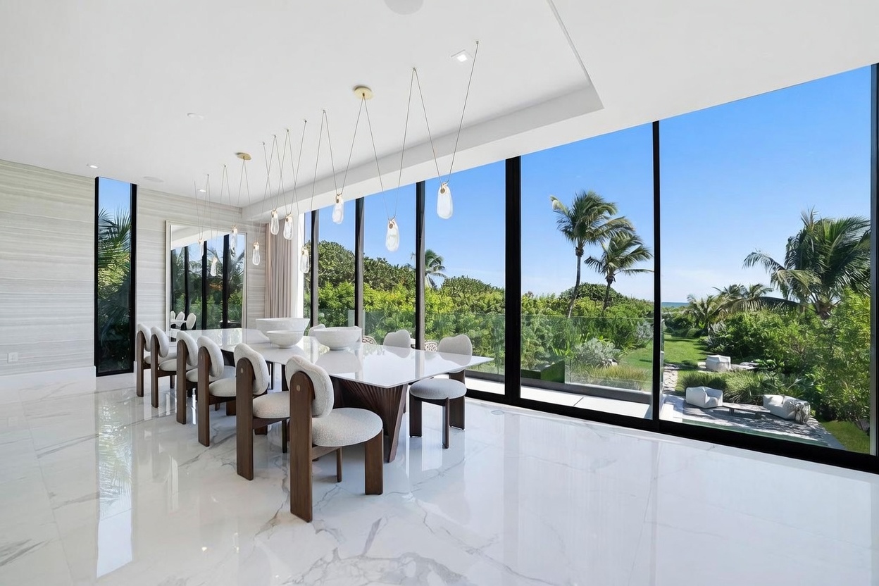 This elegant dining room features a long, glossy white table surrounded by modern chairs with dark wood frames and light-colored upholstery. The space is defined by expansive floor-to-ceiling windows that offer a panoramic view of the lush green landscape and tropical trees outside. A series of unique, organic-shaped pendant lights hang above the table, adding a sophisticated touch to the bright, open-concept interior with polished stone flooring.
