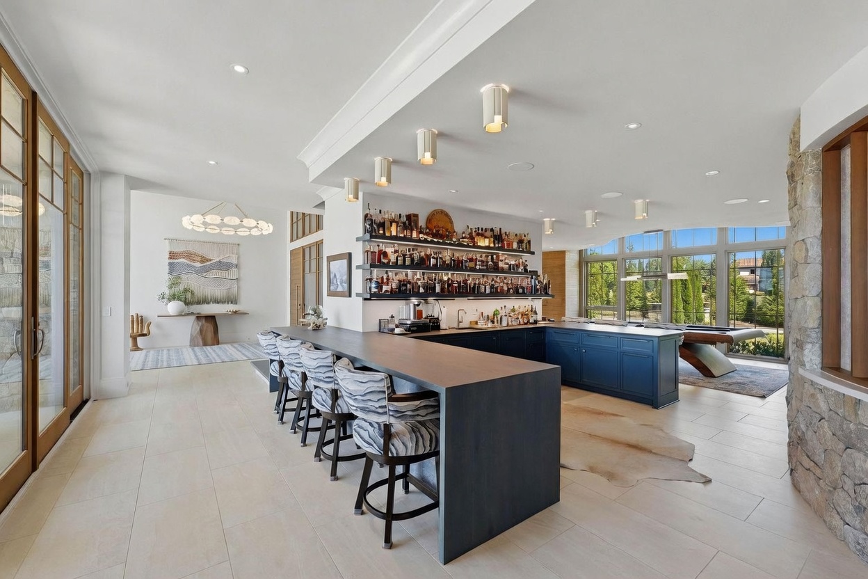 This expansive entertainment space features a large, dark-toned bar with multiple stools and a well-stocked back bar with open shelving. To the right, a pool table sits near large windows that provide a view of the outdoor landscape, while a textured wall adds a rustic element to the room. The open-concept layout includes a seating area with a console table and a decorative wall hanging, creating a sophisticated atmosphere for hosting guests.