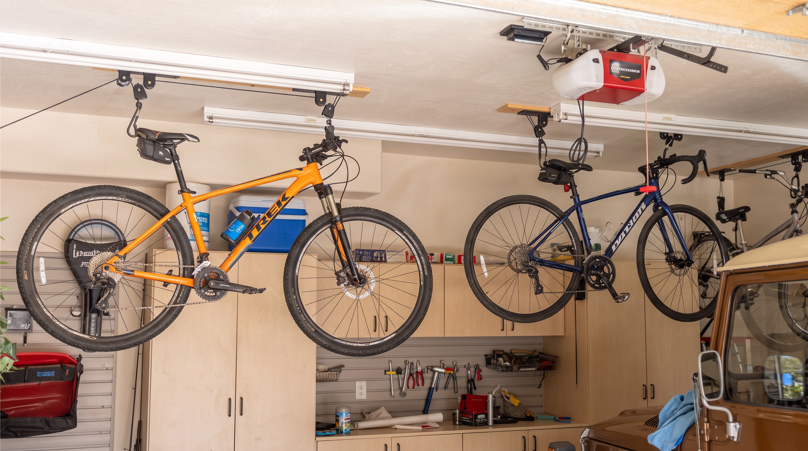 This interior view of a garage showcases efficient storage solutions, including ceiling-mounted pulley systems for hanging bicycles and custom light-wood cabinetry. The space keeps an organized aesthetic with wall-mounted tool racks, providing a tidy and functional workspace. A portion of a vintage vehicle visible in the corner adds character to this well-maintained and practical residential garage.