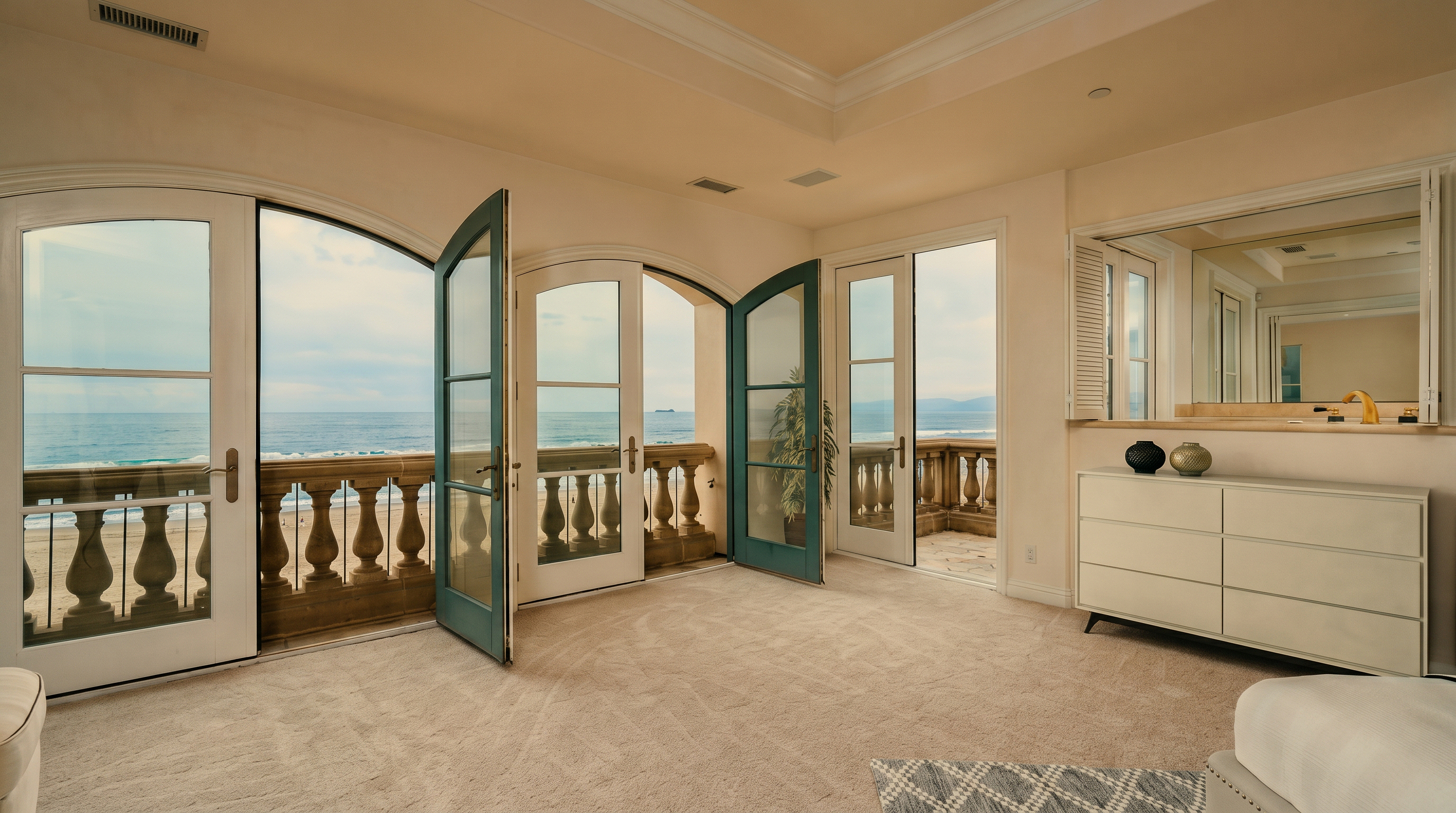 This bright and airy primary bedroom features expansive glass doors that open onto a private balcony with stunning ocean views. The room is finished with neutral carpeting and a white dresser, creating a serene and minimalist atmosphere. The architectural design emphasizes the connection between the indoor living space and the coastal scenery outside.