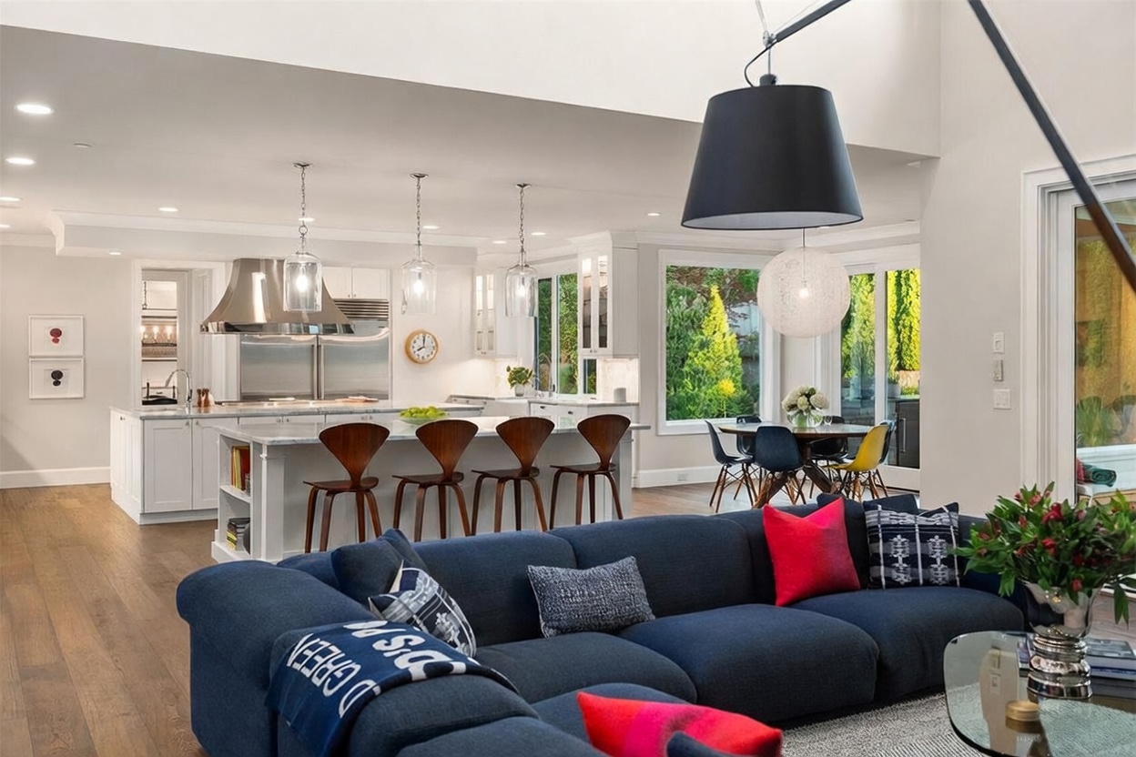 This open-concept living space features a large, deep blue sectional sofa in the foreground, leading into a bright, modern kitchen and dining area. The kitchen is equipped with white cabinetry, a large central island with bar seating, and stainless steel appliances, while the adjacent dining area is illuminated by a large pendant light. Large windows provide ample natural light and views of the outdoor greenery, creating an airy and inviting atmosphere.