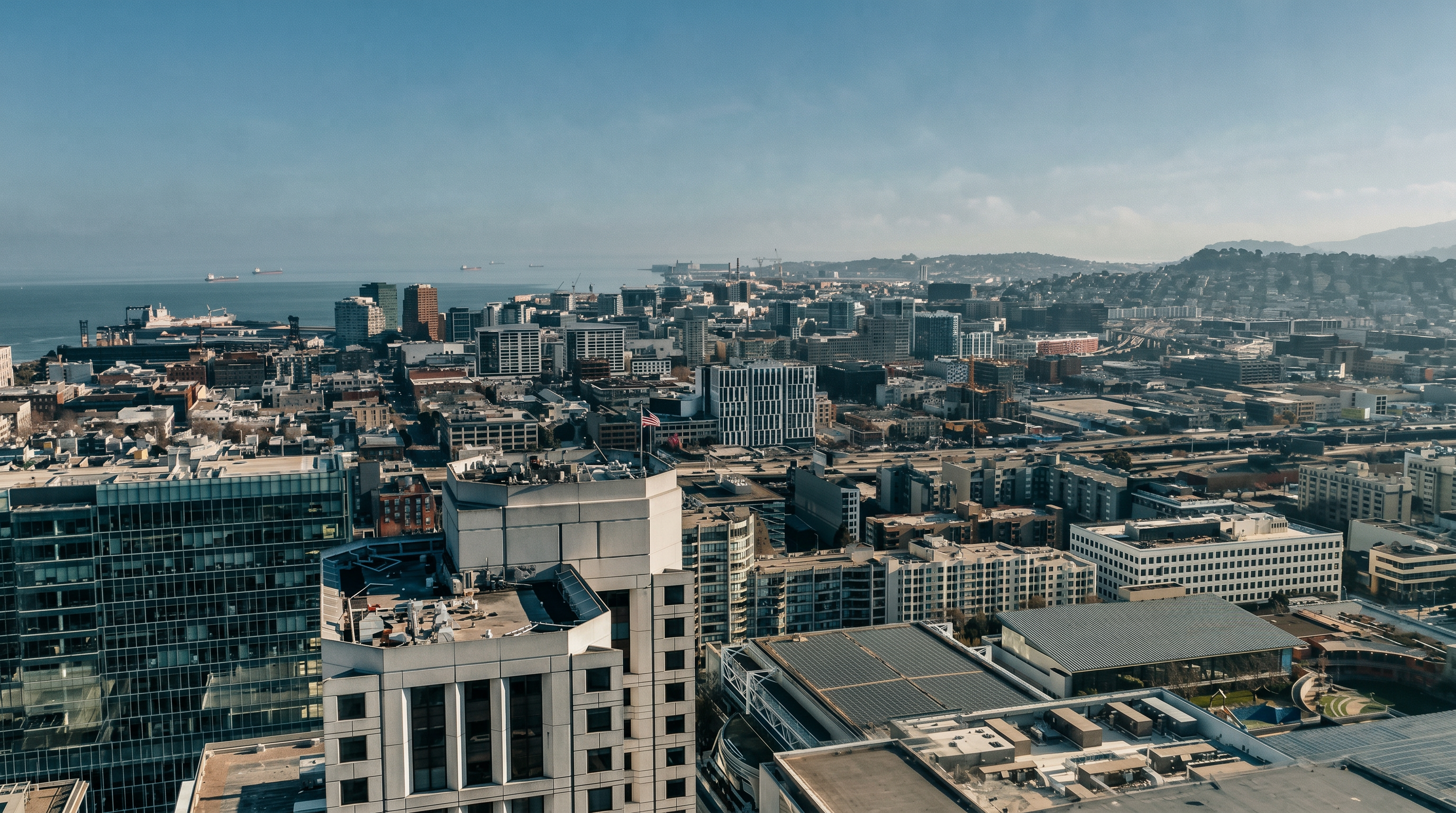 This high-altitude aerial view captures a dense urban cityscape, showcasing a mix of modern high-rise buildings and industrial structures stretching toward the distant coastline. The perspective highlights the architectural density of the area, with a prominent foreground building featuring a flat roof and a small American flag. The clear sky and expansive horizon provide a sense of scale and metropolitan connectivity.
