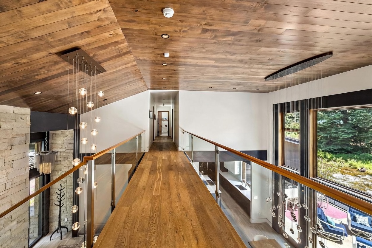 This perspective captures a dramatic bridge-style hallway that overlooks the lower level of the home, featuring a warm wood-paneled ceiling and matching wood flooring. A glass railing with a wood handrail provides an open, airy feel, while two elaborate, cascading light fixtures with spherical glass pendants flank the walkway. The space connects to a bright, minimalist hallway leading to a distant doorway, creating a sophisticated and modern architectural transition.