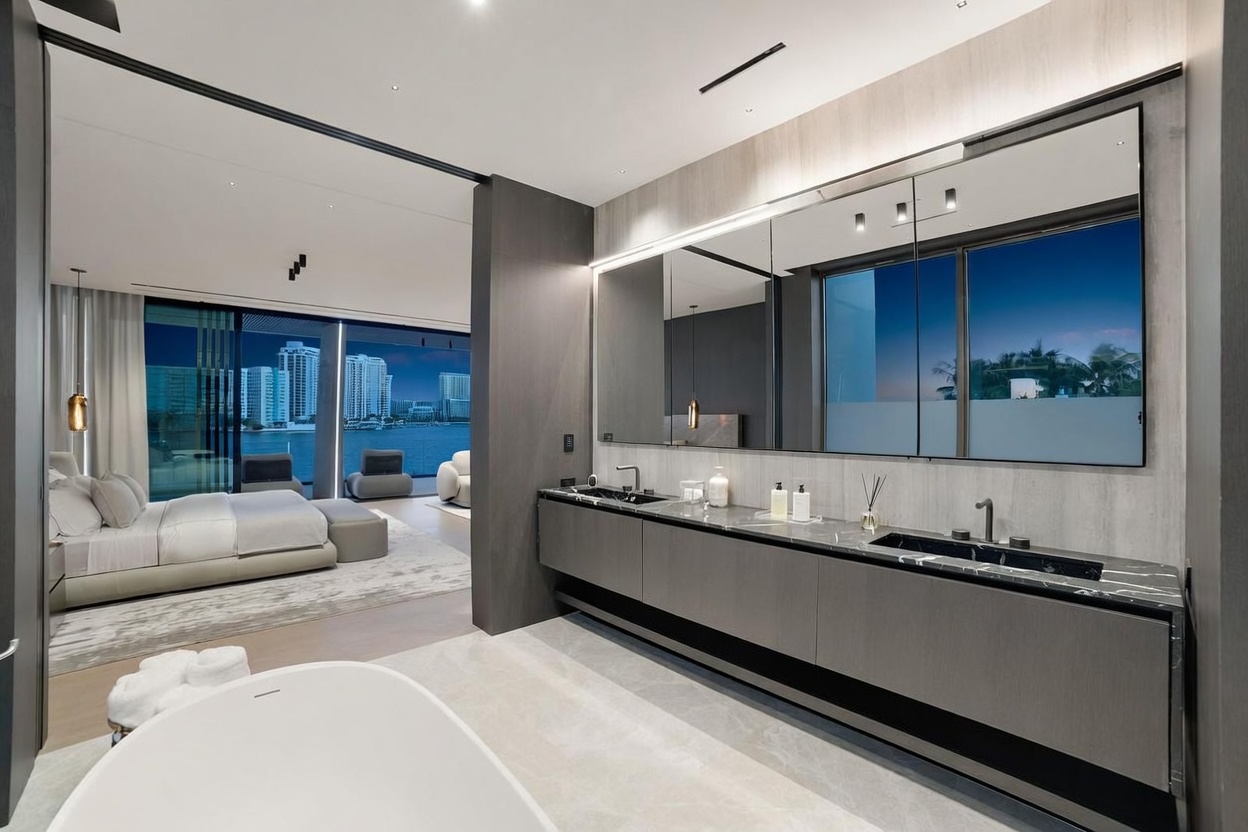 This luxurious primary bathroom features a sleek, dark-toned vanity with dual integrated sinks and a long, expansive mirror that reflects the adjacent bedroom. A freestanding white soaking tub sits in the foreground, while the open-concept design allows for a seamless transition into the bedroom, which boasts large windows overlooking a waterfront cityscape. The space is defined by its modern, minimalist aesthetic, high-end finishes, and sophisticated lighting design.