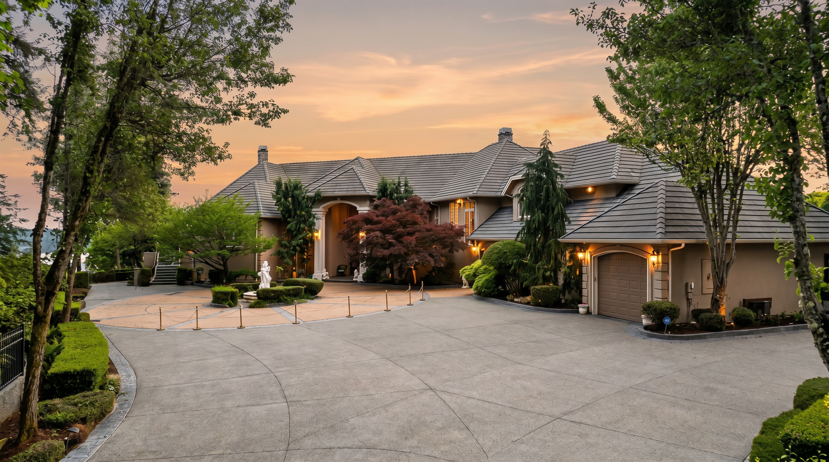 This grand estate features a sprawling concrete driveway leading to a multi-car garage and a prominent arched entryway. The home is characterized by its complex roofline, neutral-toned stucco exterior, and mature landscaping, including a striking Japanese maple tree in the circular courtyard. The perspective is a wide-angle shot from the driveway, capturing the impressive scale and curb appeal of the property under a clear blue sky.