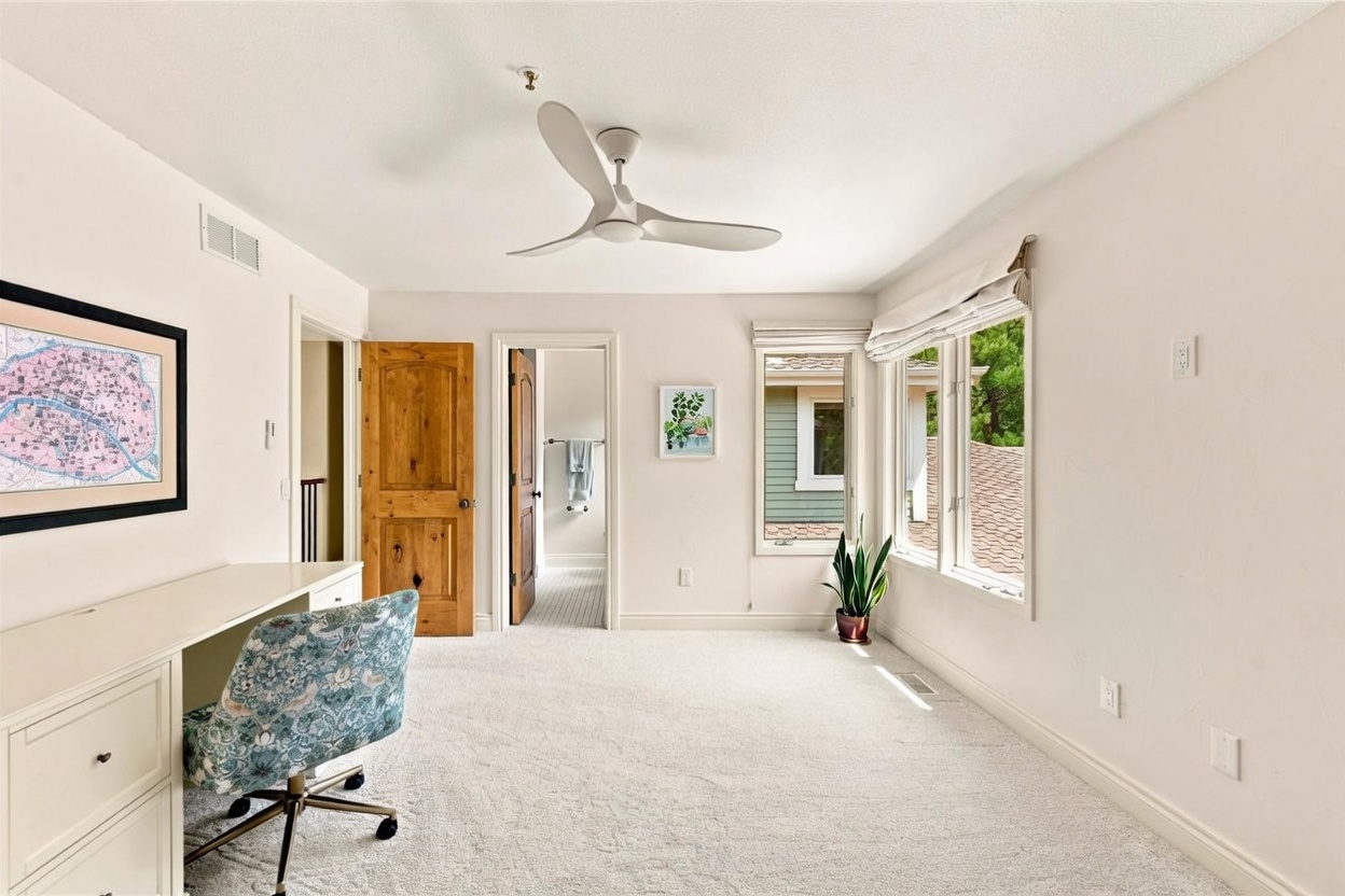 This bright and airy home office features neutral walls, plush carpeting, and a modern white desk paired with a patterned upholstered chair. A three-blade ceiling fan provides air circulation, while large windows allow for ample natural light and a view of the outdoors. The room includes a wooden door leading to an adjacent bathroom, creating a functional and well-lit workspace.