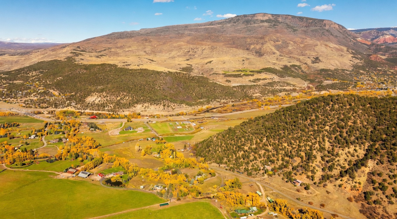 This expansive aerial view captures a vast rural landscape characterized by rolling hills, golden autumn foliage, and a sprawling valley floor. The scene features a mix of open pastures, scattered residential properties, and a winding road, all set against the backdrop of a massive, arid mountain range. The perspective provides a comprehensive look at the property's setting, emphasizing the quiet, natural beauty and the significant scale of the surrounding terrain.