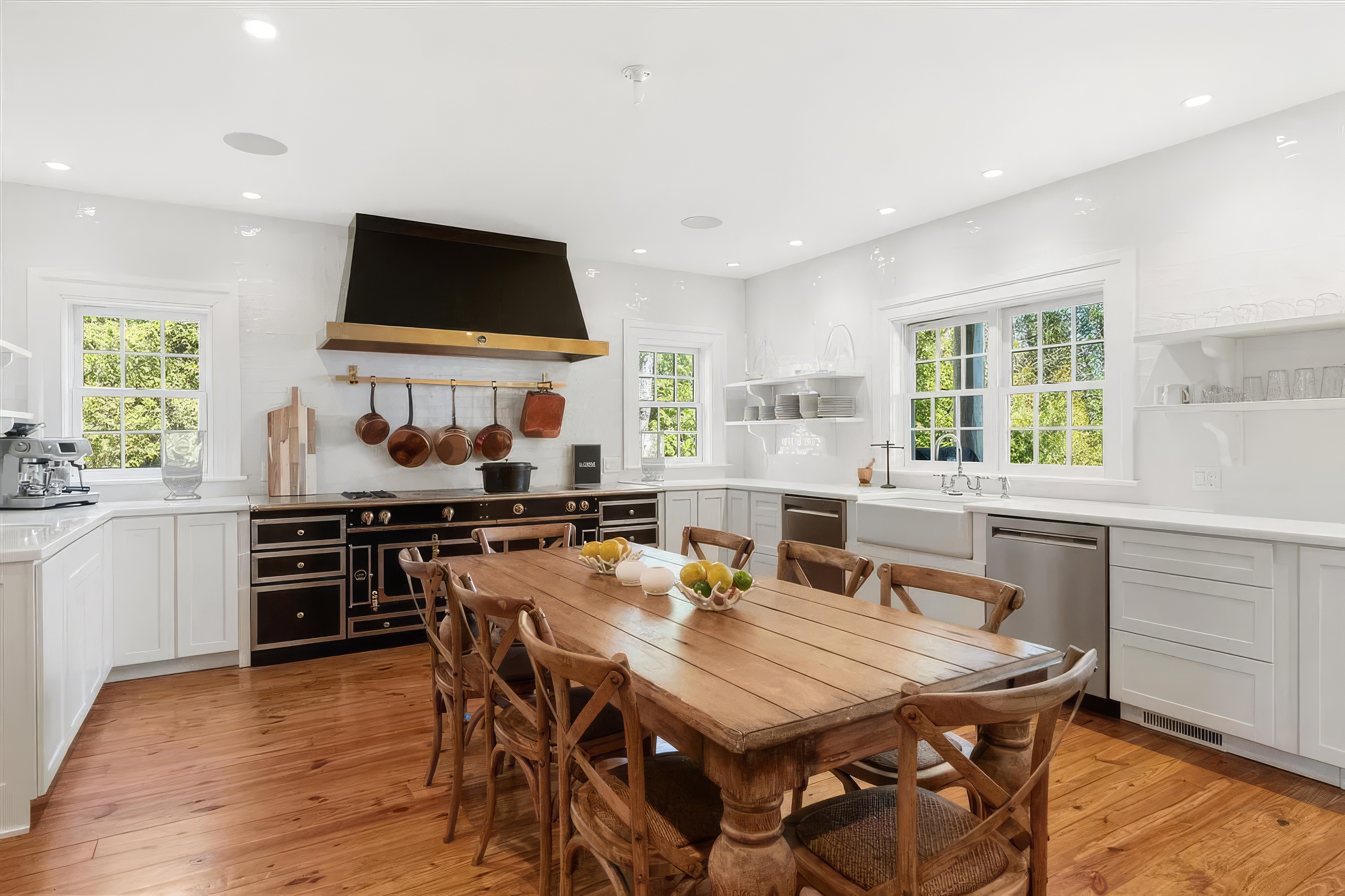 This bright and spacious kitchen features a large, rustic wooden dining table at its center, surrounded by cross-back chairs. The room is anchored by a professional-style black range with a matching hood, complemented by white cabinetry and light-colored countertops. Large windows allow natural light to fill the space, highlighting the warm wood flooring and creating an inviting, farmhouse-inspired atmosphere.