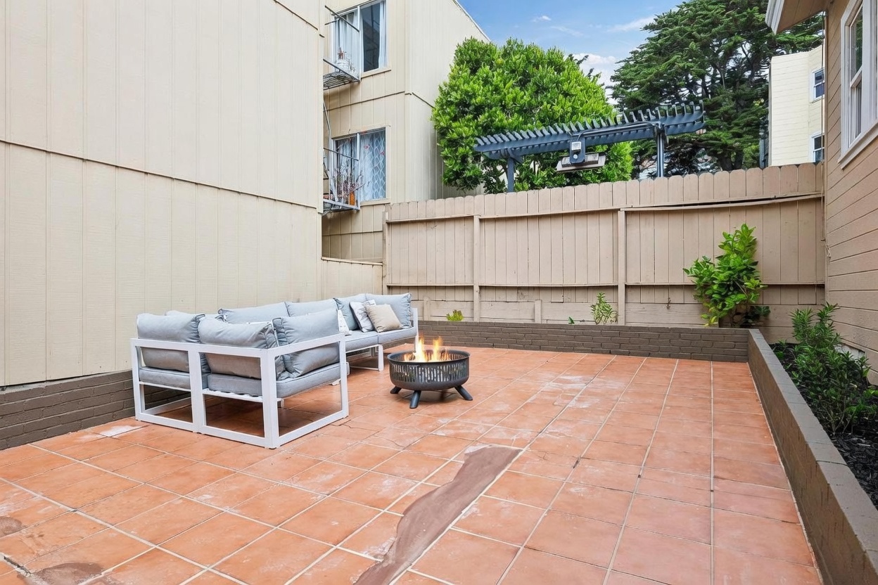This outdoor patio space features terracotta-toned tiling and a low brick retaining wall that separates the seating area from a small garden bed. A modern white-framed sectional sofa with light gray cushions is arranged around a circular metal fire pit, creating a cozy gathering spot. The area is enclosed by a tall wooden privacy fence, with a pergola structure visible in the background against a backdrop of mature trees.
