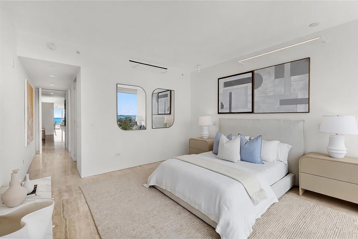 This bright and airy primary bedroom features a neutral color palette with soft blue accent pillows on a plush bed. The room is furnished with matching light-toned nightstands, elegant table lamps, and a pair of modern mirrors reflecting a view of the outdoors. A hallway leads away from the bedroom, showcasing a clean, minimalist aesthetic with light-colored flooring throughout.