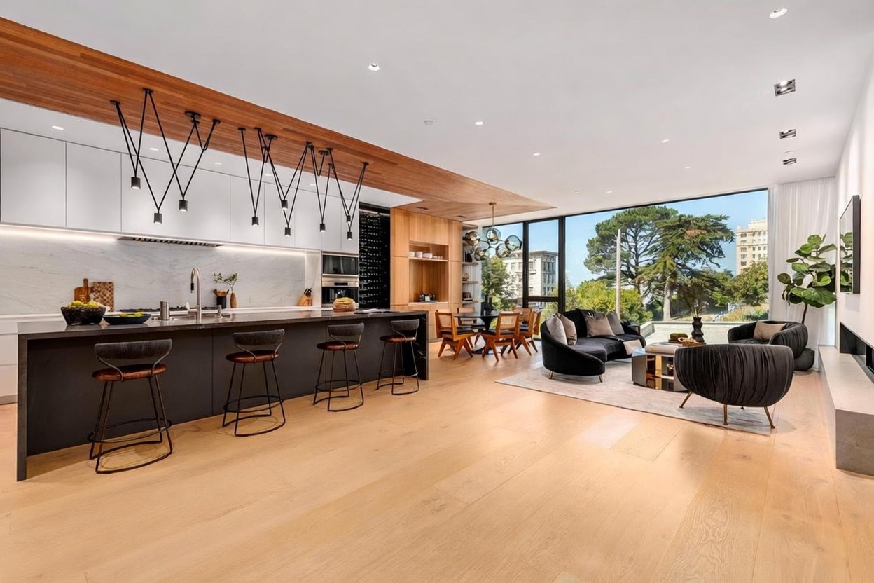 This expansive, open-concept living space seamlessly integrates a modern kitchen with a sophisticated lounge area. The kitchen features a long, dark island with bar seating, light wood cabinetry, and a striking row of pendant lights, while the adjacent living area is bathed in natural light from large windows overlooking mature trees. The design aesthetic is sleek and contemporary, characterized by clean lines, a warm wood-paneled ceiling, and a neutral color palette.