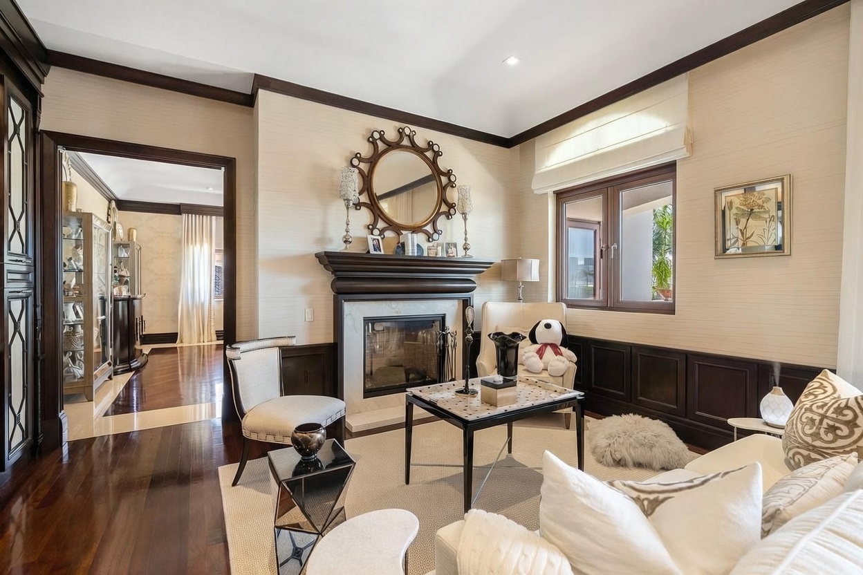 This elegant living room features a sophisticated fireplace as its focal point, adorned with a decorative mirror and dark wood trim. The space is furnished with a plush cream armchair, a modern coffee table, and a comfortable sofa, all set against a backdrop of textured wallpaper and dark wood wainscoting. The room exudes a refined, classic atmosphere with warm lighting and a seamless transition into an adjacent hallway.