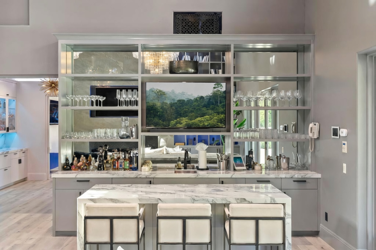 This interior shot showcases a stylish kitchen bar area, complete with a marble countertop island and three cushioned stools. The bar features a mirrored backdrop with shelving displaying glassware and liquor bottles, centered around a television. The overall design is modern and sophisticated, creating an inviting space for entertaining.
