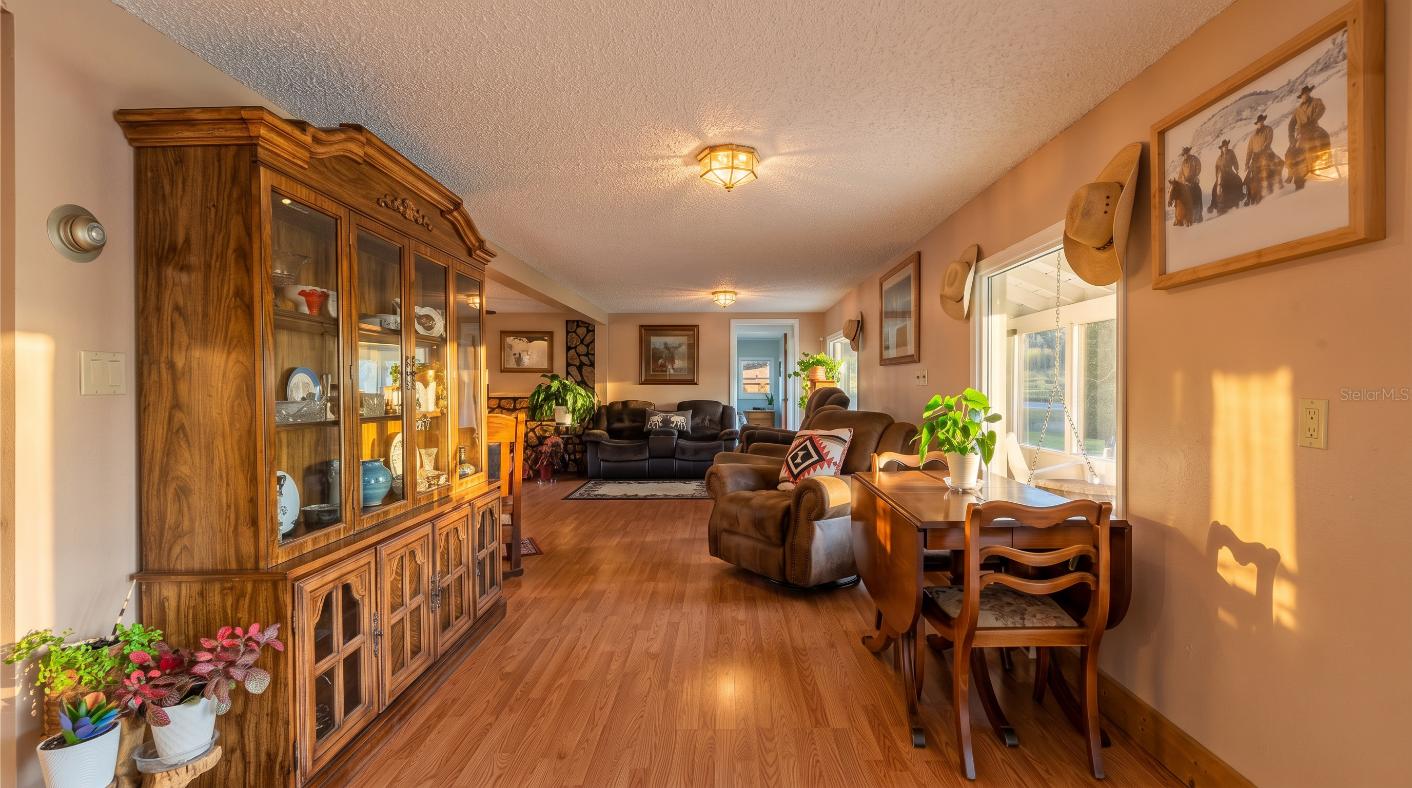 This open-concept living area features warm wood-look flooring and a traditional, cozy aesthetic. A large, ornate wooden hutch stands prominently on the left, while a dining set sits near a window on the right, leading into a spacious living room with plush seating in the background. The space is decorated with western-themed artwork and cowboy hats, creating a rustic and inviting atmosphere.