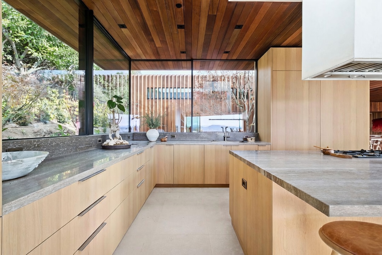 This modern, minimalist kitchen features warm wood cabinetry paired with sleek, grey stone countertops and a matching backsplash. A large floor-to-ceiling window provides an expansive view of the lush outdoor greenery, seamlessly blending the interior with nature. The space is defined by its clean lines, high-end finishes, and a bright, airy atmosphere that emphasizes contemporary luxury.
