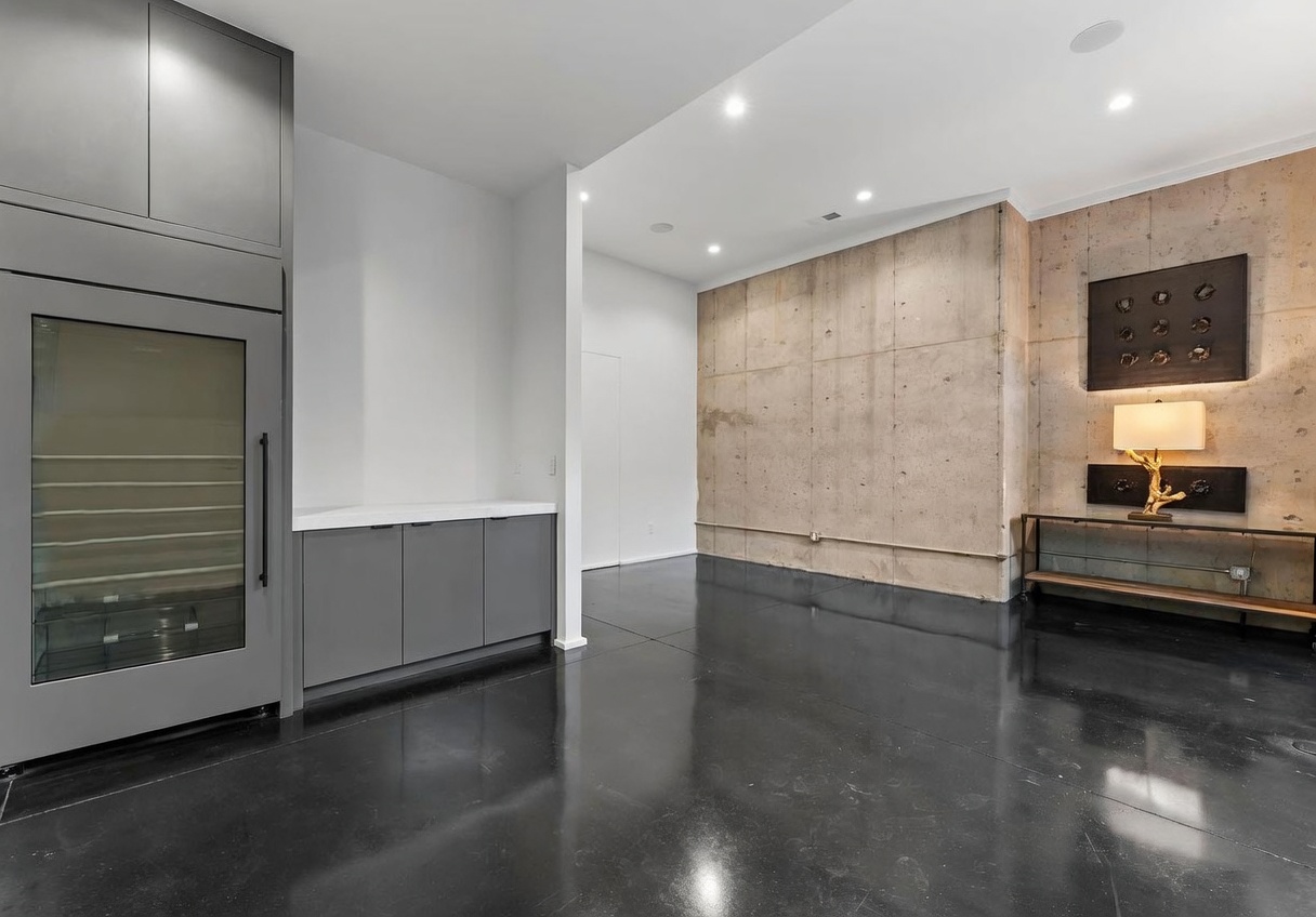 This modern, industrial-style space features a built-in wine refrigerator integrated into dark cabinetry with a light-colored countertop. The room is anchored by a prominent, textured concrete wall adorned with a decorative art piece and a warm-toned lamp on a metal console table. The polished dark flooring reflects the recessed ceiling lights, creating a sleek and sophisticated atmosphere.
