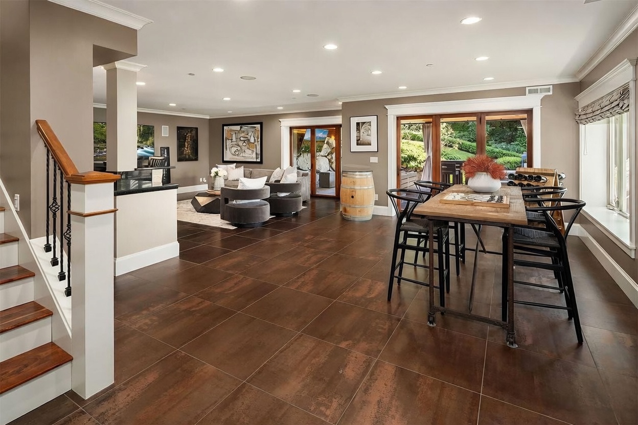 This open-concept living and dining area features large-format, dark-toned tile flooring and a neutral color palette. The space includes a dining area with a rustic wooden table and black chairs, transitioning into a comfortable living room with plush seating and modern artwork. Large glass doors provide a seamless connection to the outdoor space, while recessed lighting and a staircase with a wooden handrail add architectural interest.