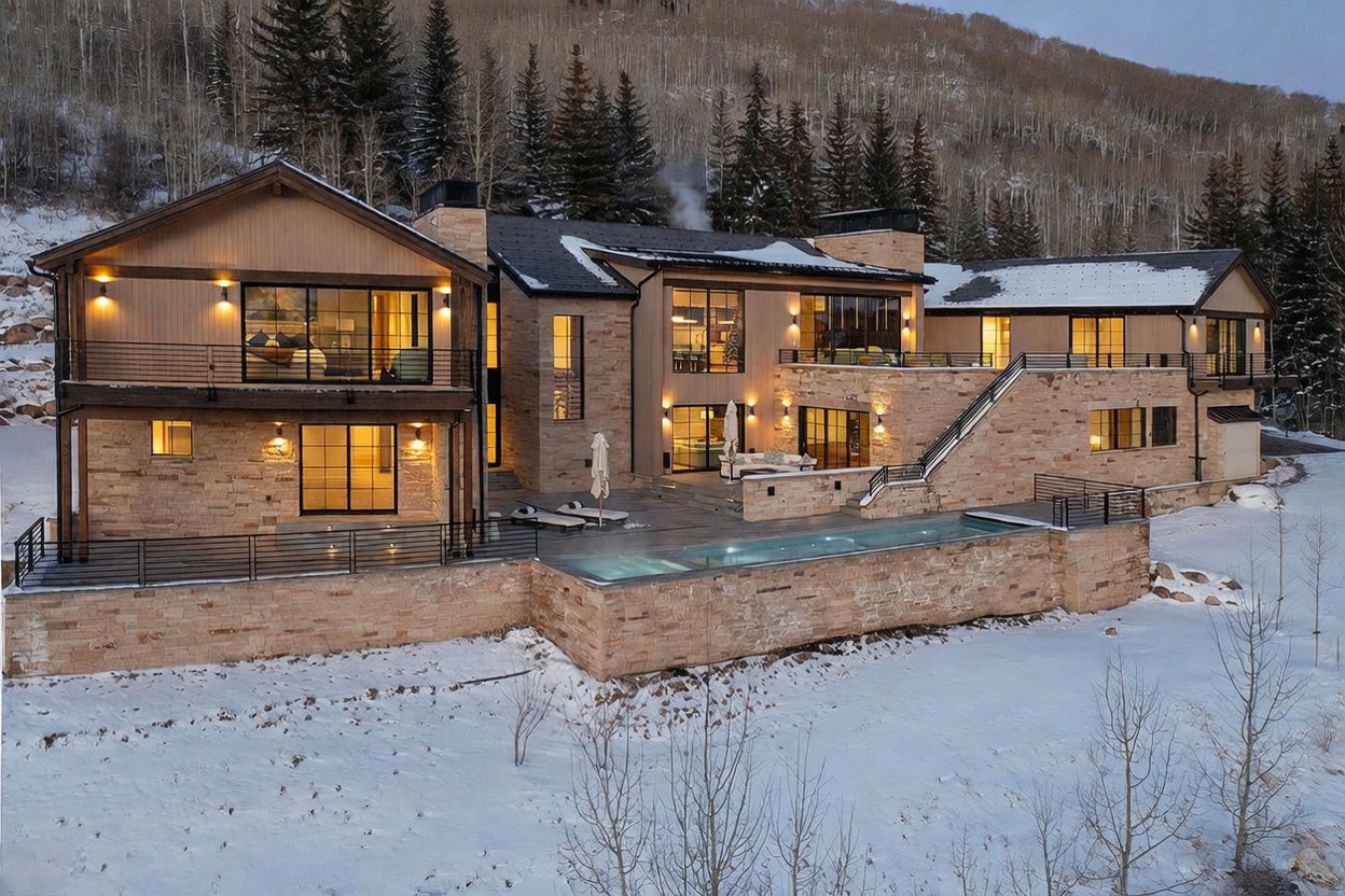 This expansive mountain estate features a sophisticated blend of natural stone and warm wood siding, set against a backdrop of snow-covered pine trees. The rear elevation showcases a multi-level design with a prominent infinity-edge pool, a spacious stone patio, and large windows that offer a seamless connection between the indoor and outdoor living spaces. The cinematic perspective captures the home's architectural grandeur and its serene, elevated position within a winter landscape.