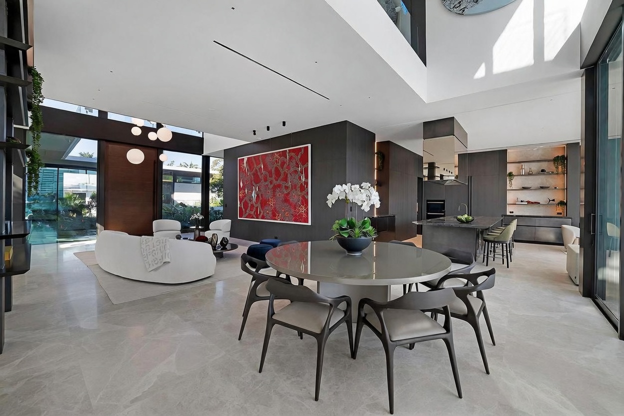 This expansive, open-concept living and dining area features high ceilings and a sophisticated, modern aesthetic with neutral stone flooring throughout. The space is anchored by a large, circular dining table and a curved white sofa, complemented by a bold, abstract red artwork on a dark accent wall. Large windows provide ample natural light and views of the outdoor greenery, creating a seamless transition between indoor and outdoor living.