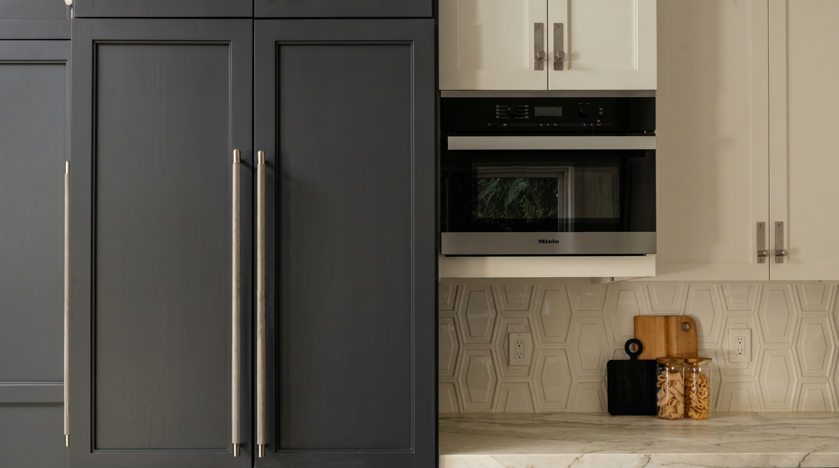 This kitchen features a clean, high-contrast aesthetic with matte dark gray cabinetry paired with white upper cabinets and a built-in Miele microwave. The countertop boasts a white marble look, accented by a modern hexagonal tile backsplash and subtle decorative wooden cutting boards. The combination of stainless steel hardware and refined finishes creates a sophisticated, modern culinary space.