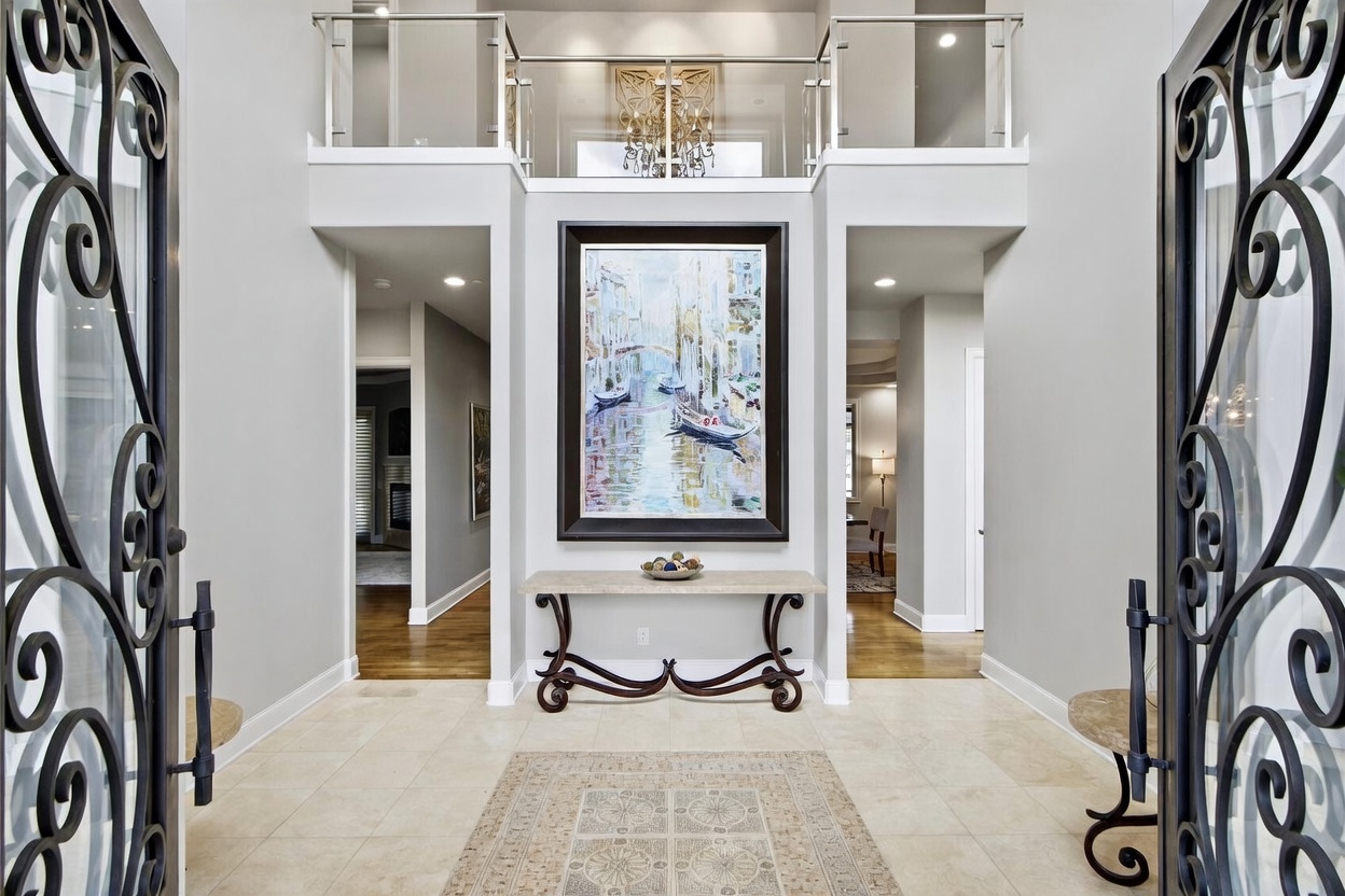 This grand entryway features a double-height ceiling and an open mezzanine with a glass railing, creating an airy and sophisticated atmosphere. A large, framed painting of a canal scene serves as a focal point above a console table, while ornate metal-framed doors frame the view into the home. The space is finished with light-toned walls and neutral tile flooring, providing a bright and welcoming transition into the residence.