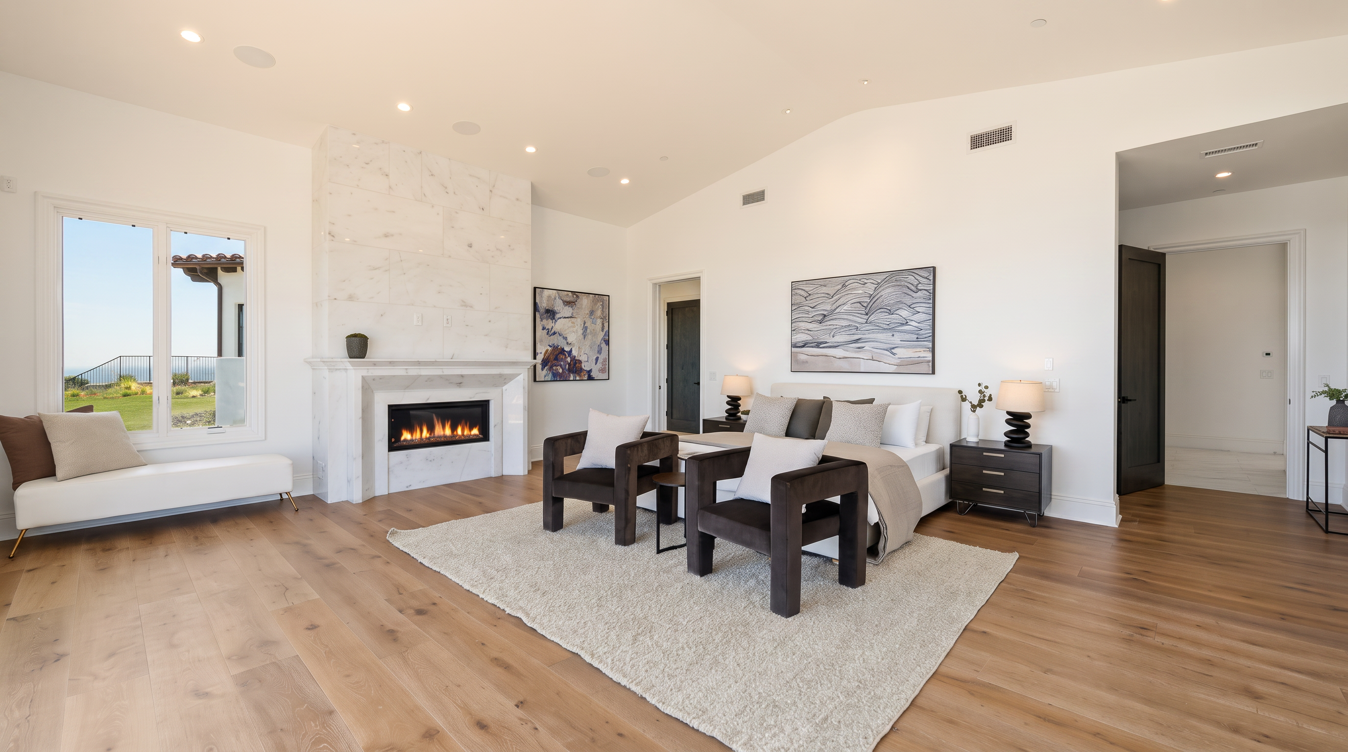 This spacious primary bedroom features light wood flooring, a neutral color palette, and a tall stone-clad chimney breast surrounding a modern fireplace. A large bed sits centrally on a textured area rug, flanked by two dark wood nightstands, while a long bench is positioned beneath a large window to the left. The room is bright and airy, with high ceilings and a minimalist aesthetic that creates a serene and luxurious atmosphere.