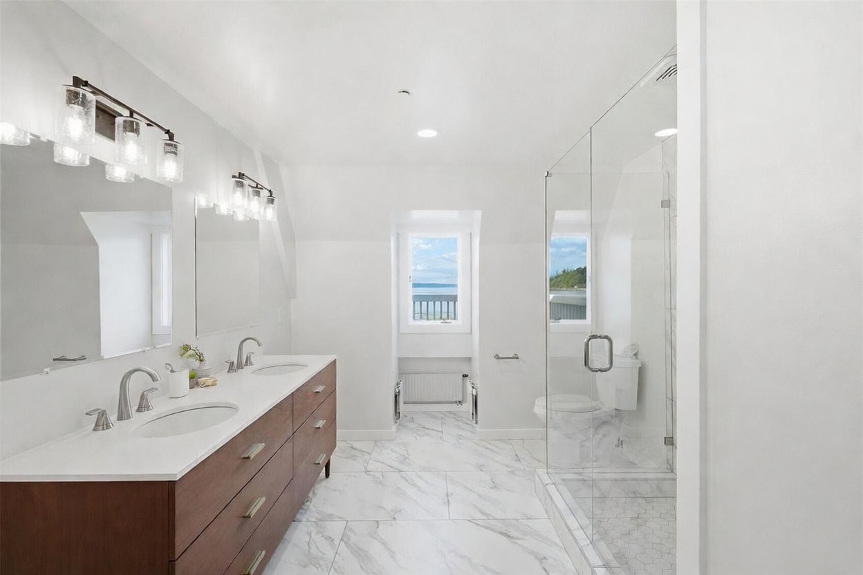 This bright, modern bathroom features a long vanity with a light-colored countertop and wood-toned cabinetry, paired with a large mirror and contemporary light fixtures. To the right, a glass-enclosed shower with patterned tile flooring adds a sleek, open feel to the space. A window at the far end provides natural light and a view of the outdoors, contributing to the clean and airy aesthetic of the room.