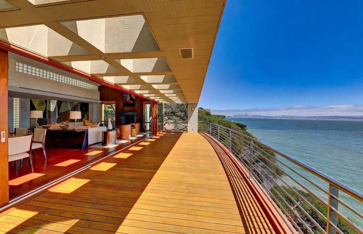 This expansive, curved wooden deck offers a panoramic view of the bay and distant city skyline, seamlessly connecting the indoor living area through large sliding glass doors. The deck features a modern metal railing and is partially sheltered by a geometric, slatted roof structure that casts rhythmic shadows across the planks. The interior space visible through the open doors includes a comfortable seating area with neutral-toned furniture, creating a sophisticated indoor-outdoor living experience.