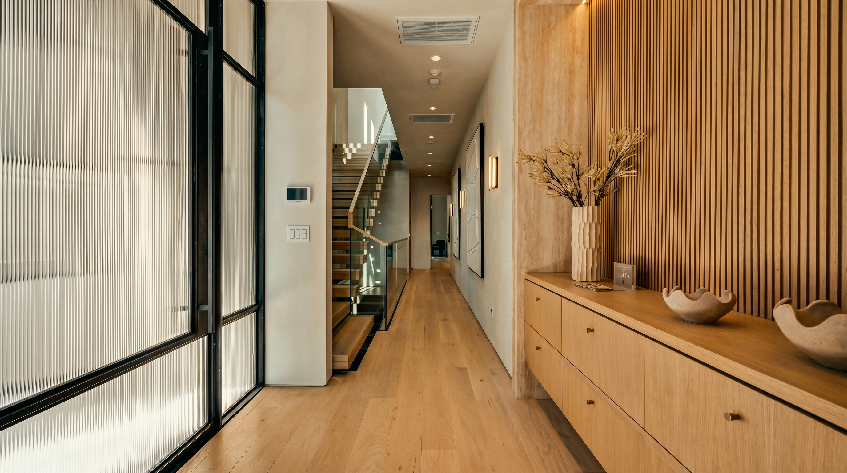 This modern hallway features light wood flooring and a striking slatted wood accent wall that adds texture and warmth to the space. A glass-railed staircase leads to the upper level, while a long, low-profile light wood console provides storage and display space. The perspective looks down the length of the corridor, highlighting the clean lines and contemporary architectural design.