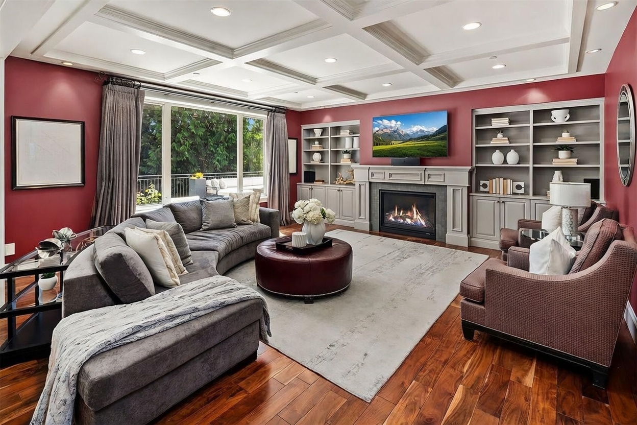 This elegant living room features deep red walls, rich wood flooring, and a coffered ceiling with recessed lighting. A large, curved sectional sofa and a leather ottoman are centered around a fireplace with a traditional mantel and a television mounted above. Built-in shelving units flank the fireplace, while large windows provide a view of the outdoor deck and greenery.