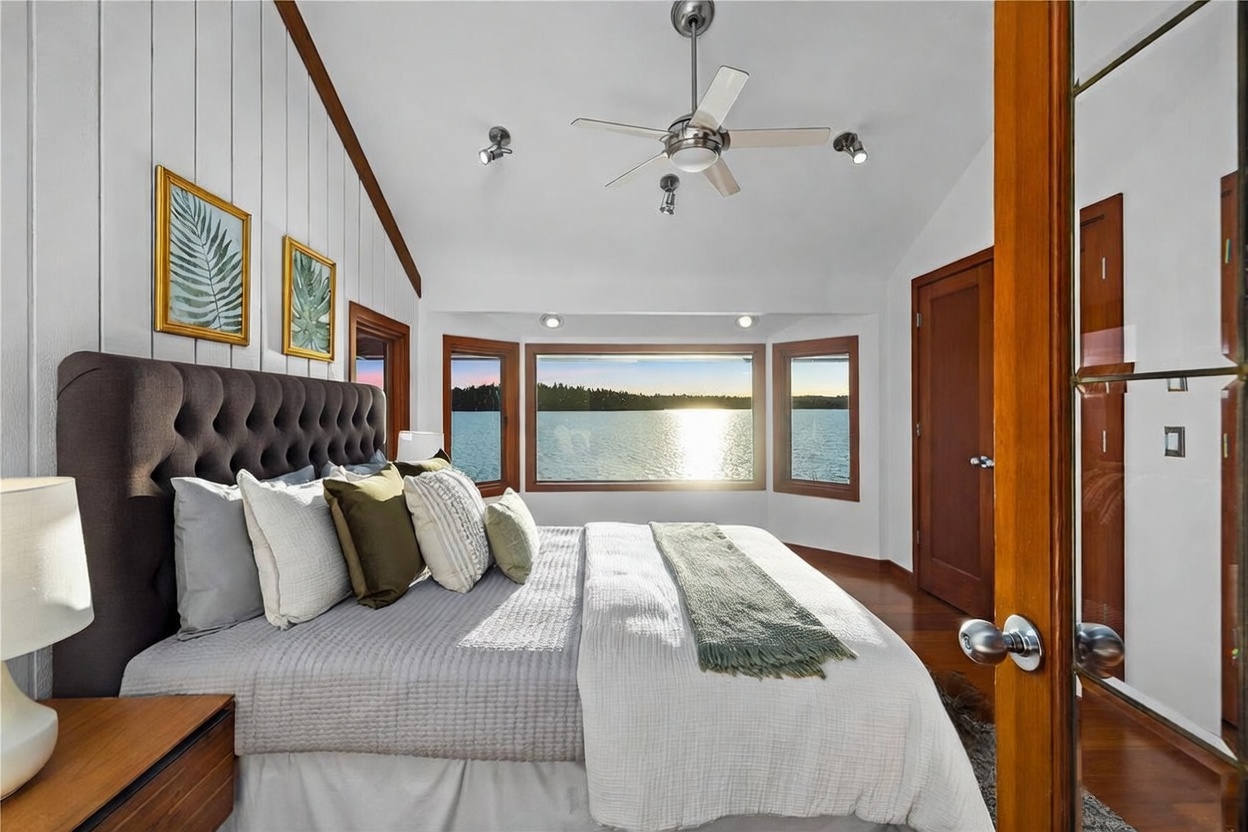 This primary bedroom features a sophisticated, modern aesthetic with a tufted dark gray headboard and a vaulted ceiling accented by wood trim. A large bay window serves as the focal point, offering a stunning, unobstructed view of a serene lake at sunset. The room is framed from a doorway perspective, highlighting the warm wood flooring and elegant, neutral-toned bedding.