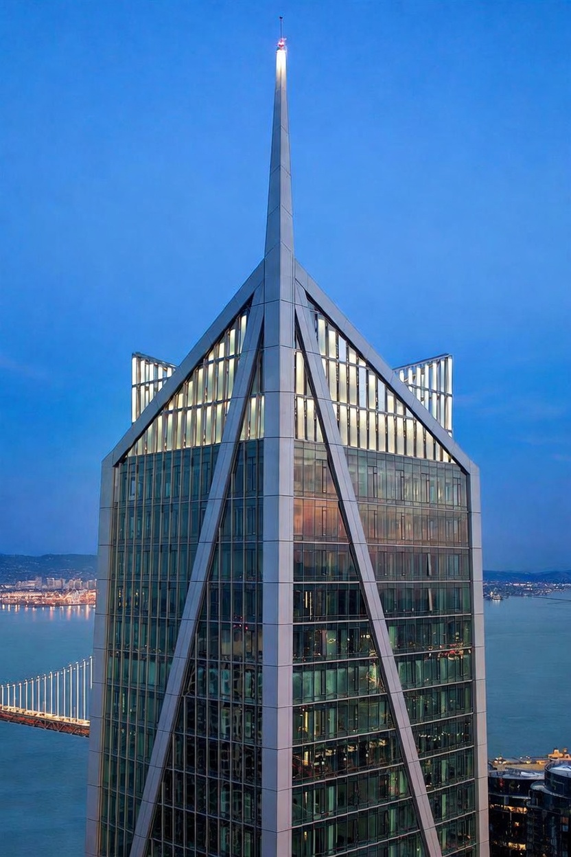 This striking aerial perspective captures the upper levels of a modern skyscraper at twilight, showcasing its unique, angular architectural design and prominent spire. The building features extensive glass paneling that reflects the soft blue sky, while the surrounding cityscape and a distant bridge are visible in the background. The composition emphasizes the building's height and sophisticated, contemporary aesthetic.