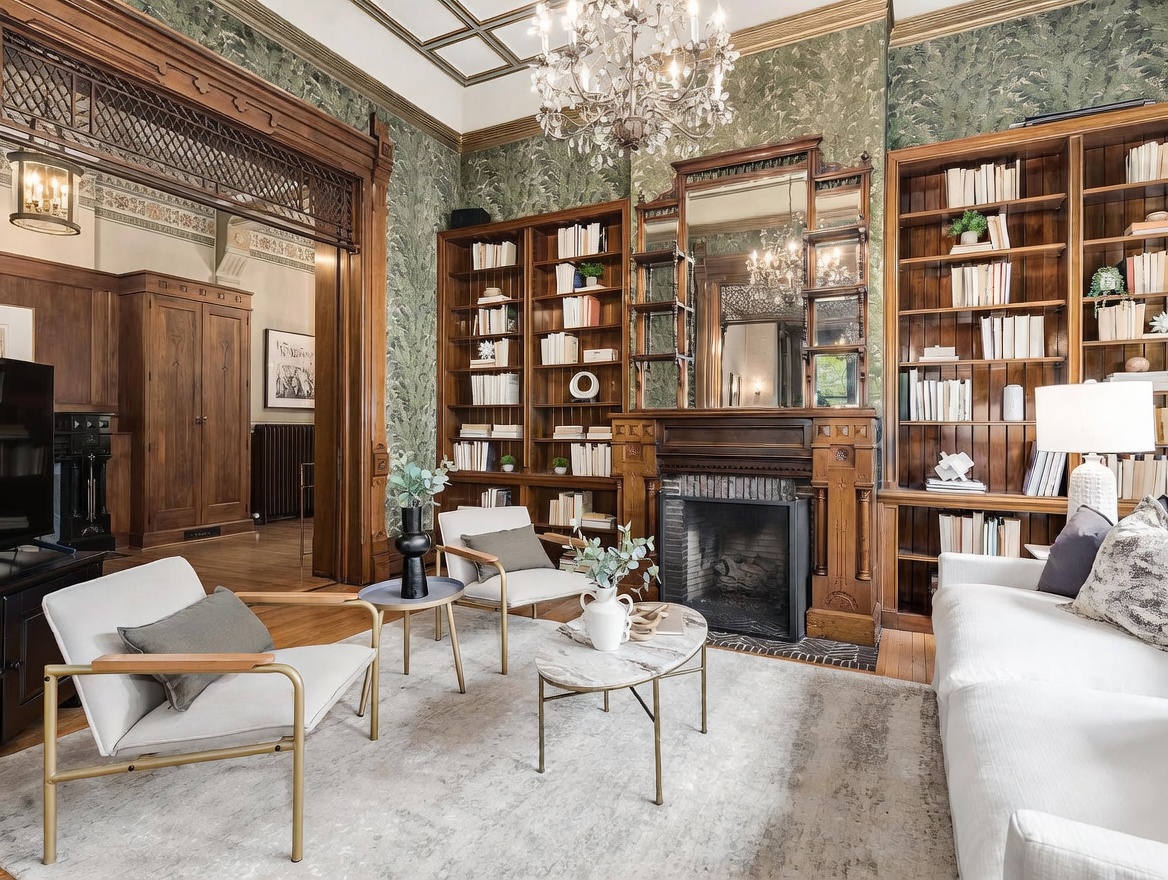 This elegant living room features rich, dark wood built-in shelving flanking a central fireplace with a decorative wood mantel and mirror above. The space is adorned with patterned green wallpaper, a crystal chandelier, and a neutral color palette for the furniture, including two modern armchairs and a sofa. The perspective is a wide-angle shot from the center of the room, showcasing the classic architectural details and the transition into an adjacent room through a large, ornate wooden archway.